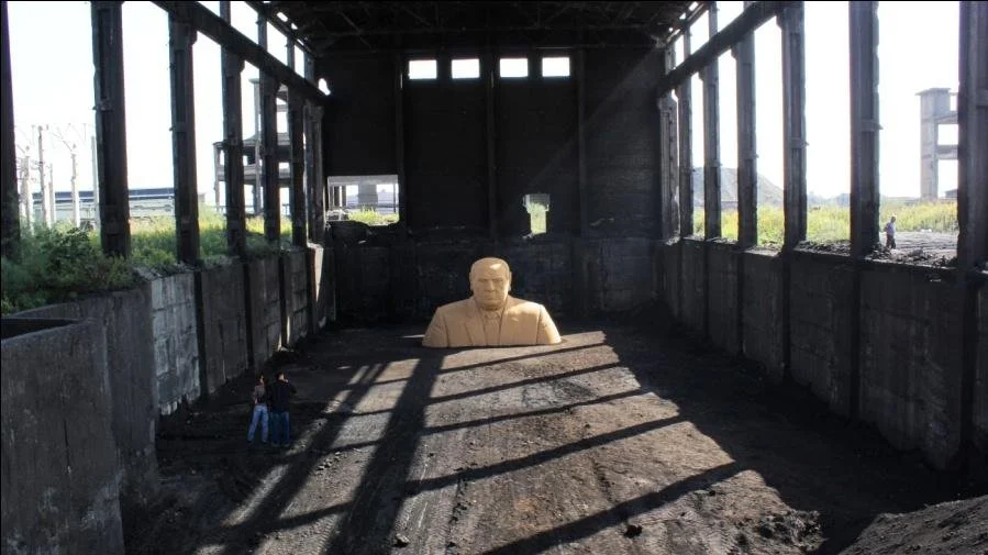A large, partially constructed or abandoned industrial building with a giant statue of a man's head and shoulders in the background. Two people are standing in front of the statue. The structure has large windows, some missing glasses, and dirt floor