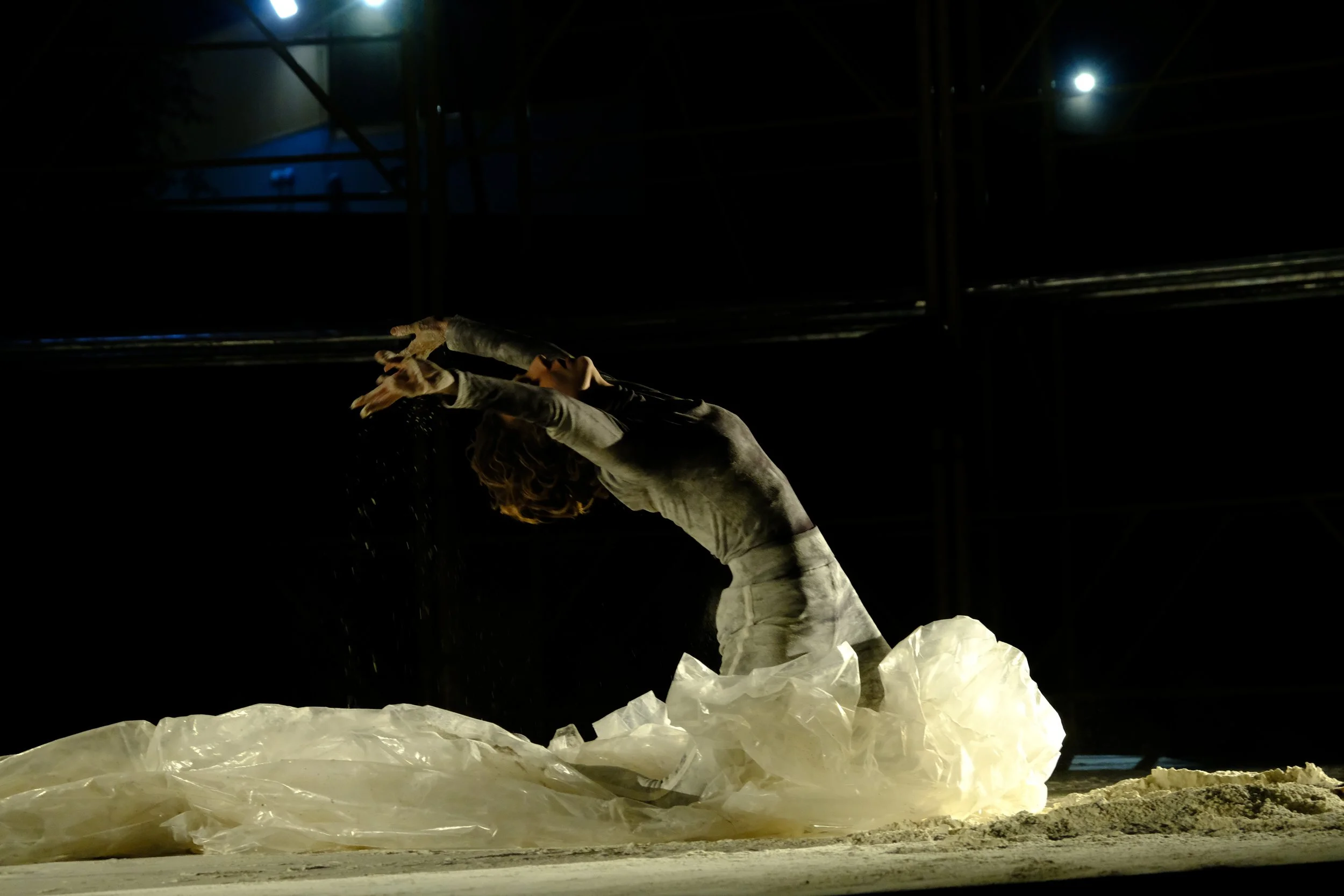 A person wearing a white outfit with curly hair stands on a illuminated rocky surface at night, with the dark sky and some bright lights in the background.
