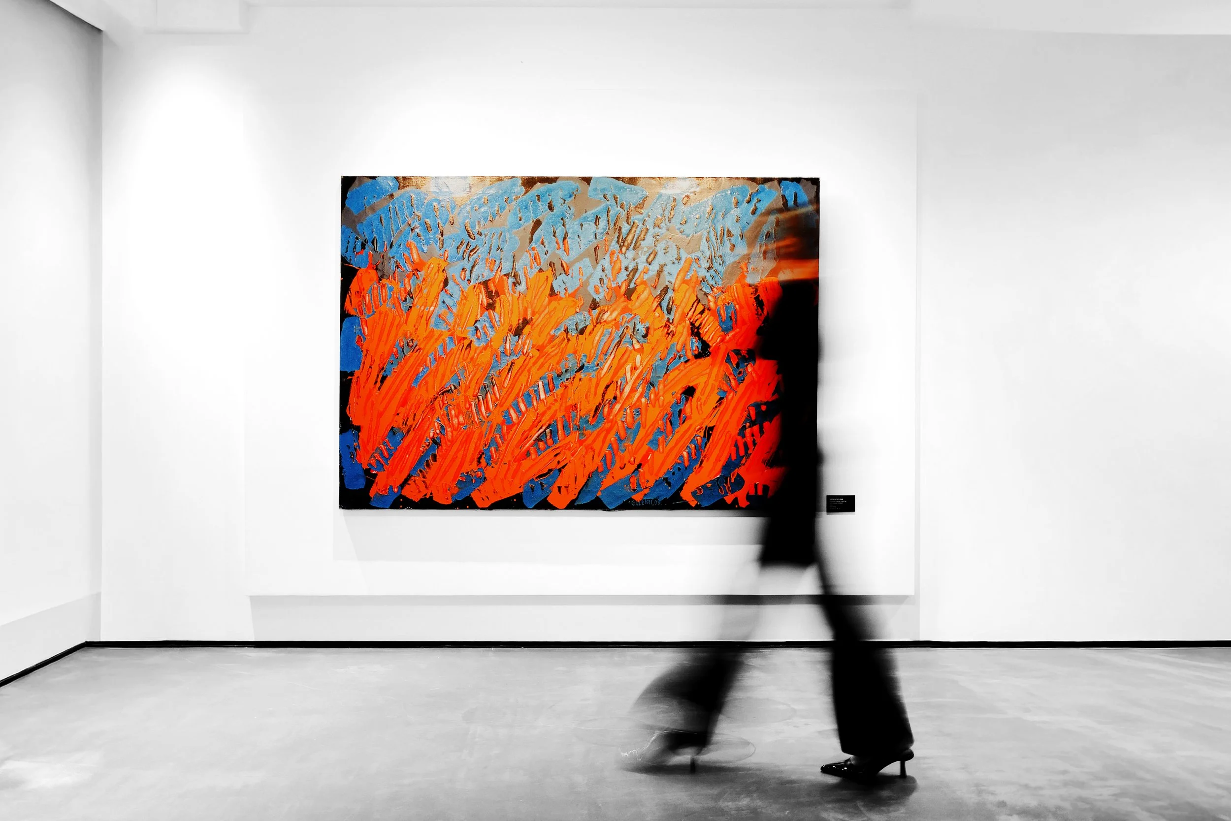 An abstract painting with vibrant orange, blue, and neutral colors displayed on a white gallery wall, with a blurred person walking in front of it.