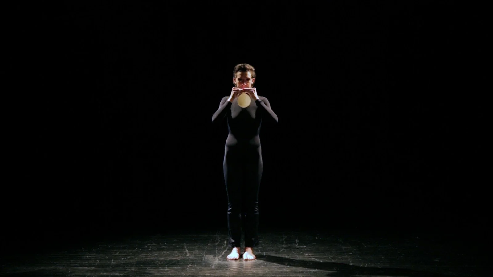A woman standing barefoot on a dark stage, playing a flute, with a focused expression, dressed in black.