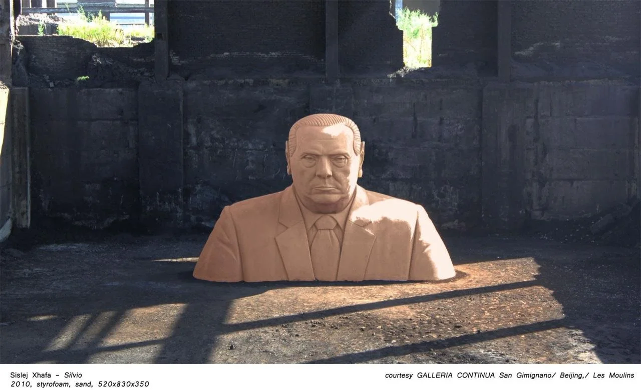 Sand sculpture of a serious-looking man in a suit with short hair, situated outdoors on a sandy surface with a blackened wall behind it.