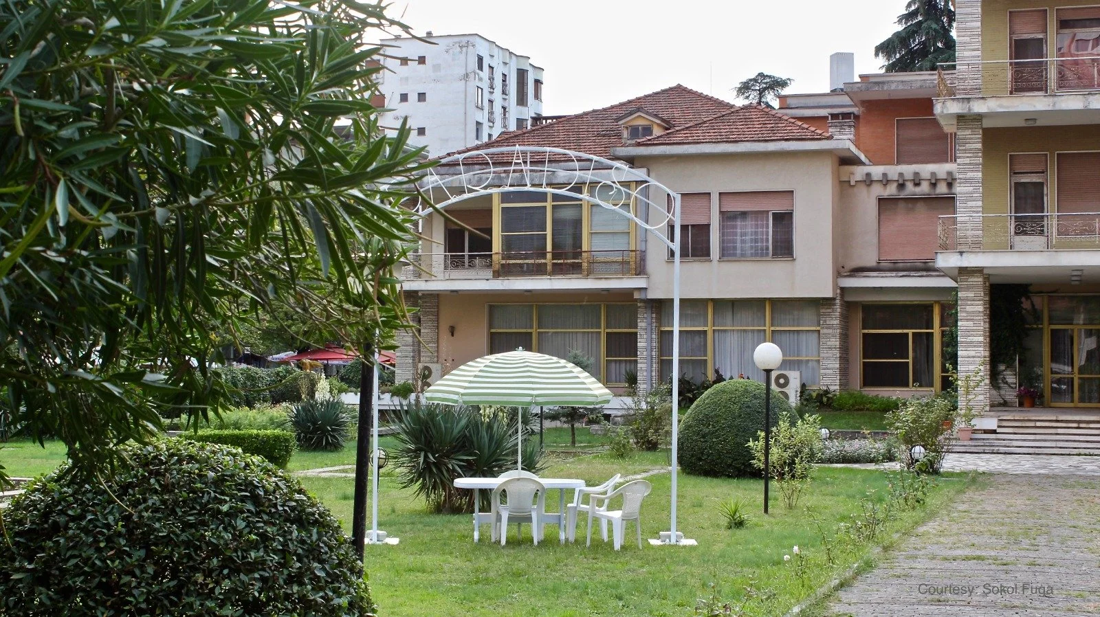 A garden area with a white patio table and four chairs under a striped umbrella, surrounded by green bushes and plants, with multi-story residential buildings in the background.