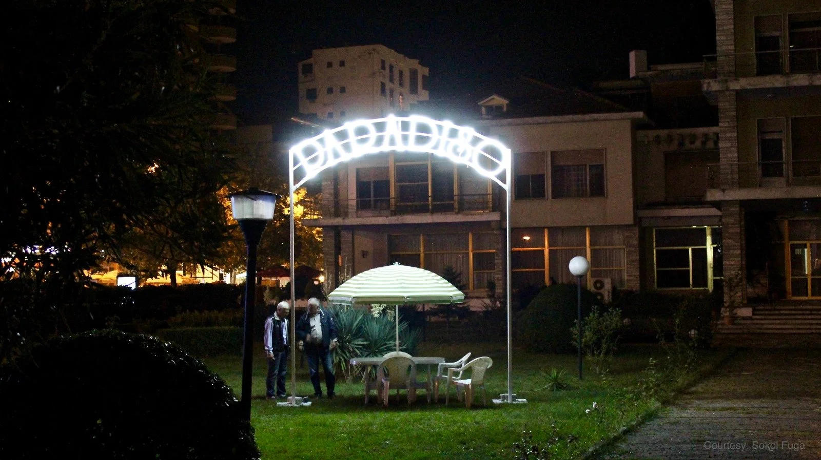 Nighttime outdoor scene with a lit-up archway sign that reads 'Badadiso', a patio table with a green and white striped umbrella, and two elderly men standing nearby on a grassy area near a building.