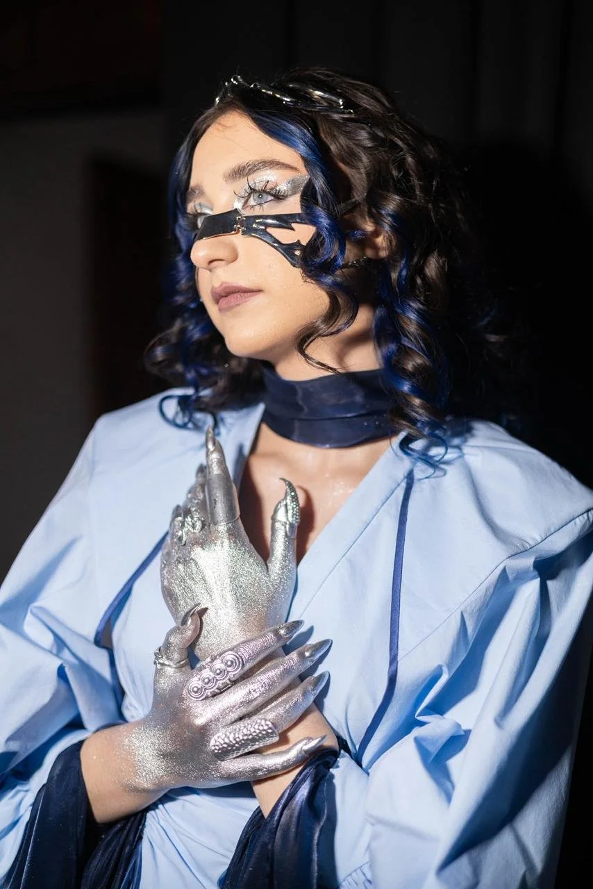 A woman with dark hair and blue streaks, wearing a black headpiece and clear glasses, poses with her hands crossed over her chest. She is dressed in a light blue garment with dark accents and has silver, metallic gloves with intricate designs. Her ma