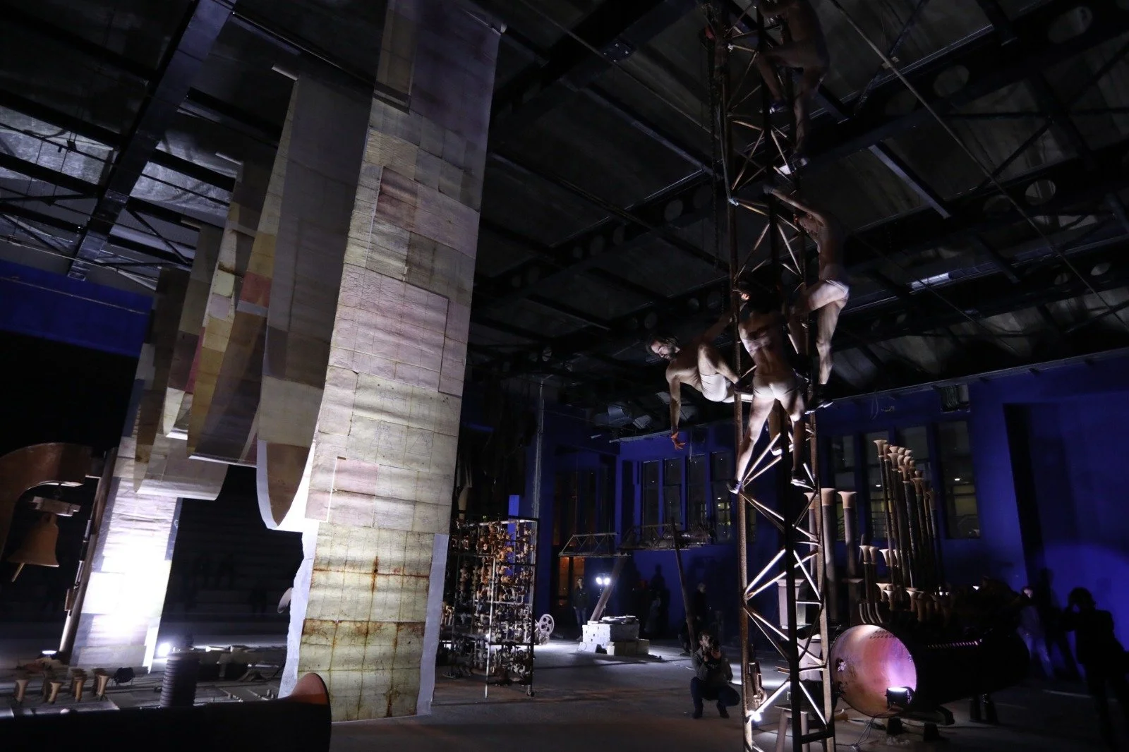 Dark indoor scene with a large vertical structure resembling a wall made of uneven, rusted metal panels. To the right, there is an artistic suspended sculpture of two human figures made of metal, intertwined and hanging from a metallic framework. The