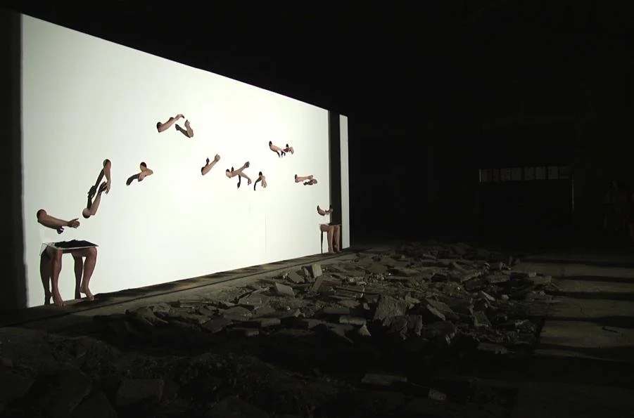 Art installation of human figures leaning forward with their heads obscured, positioned along a white wall in a dark space, with broken rubble on the floor in front of the wall.