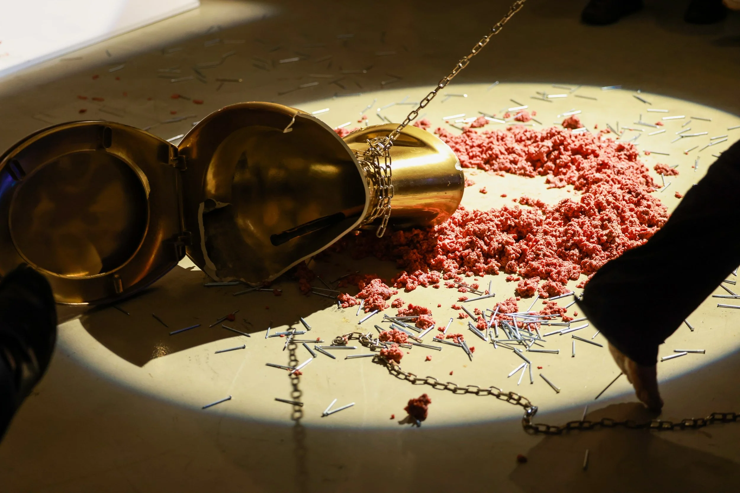 A shiny, gold-colored object that resembles a horse's head lying on the floor surrounded by scattered metal nails and pink material, with a chain attached to the object, in a dimly lit room.