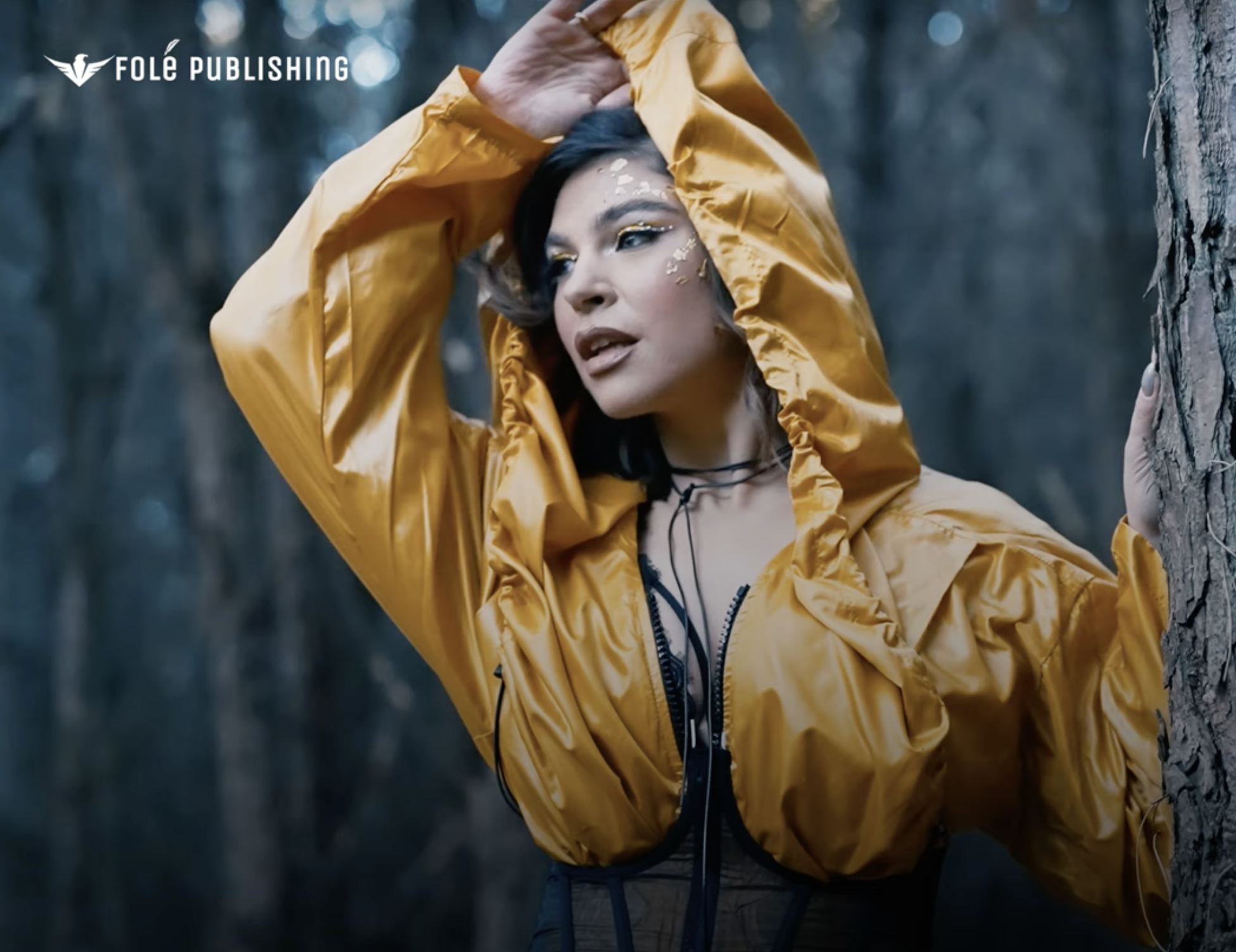 A woman wearing a shiny yellow rain jacket and black hair with makeup, including gold glitter eye makeup and decorative face embellishments, leaning against a tree in a forested area.