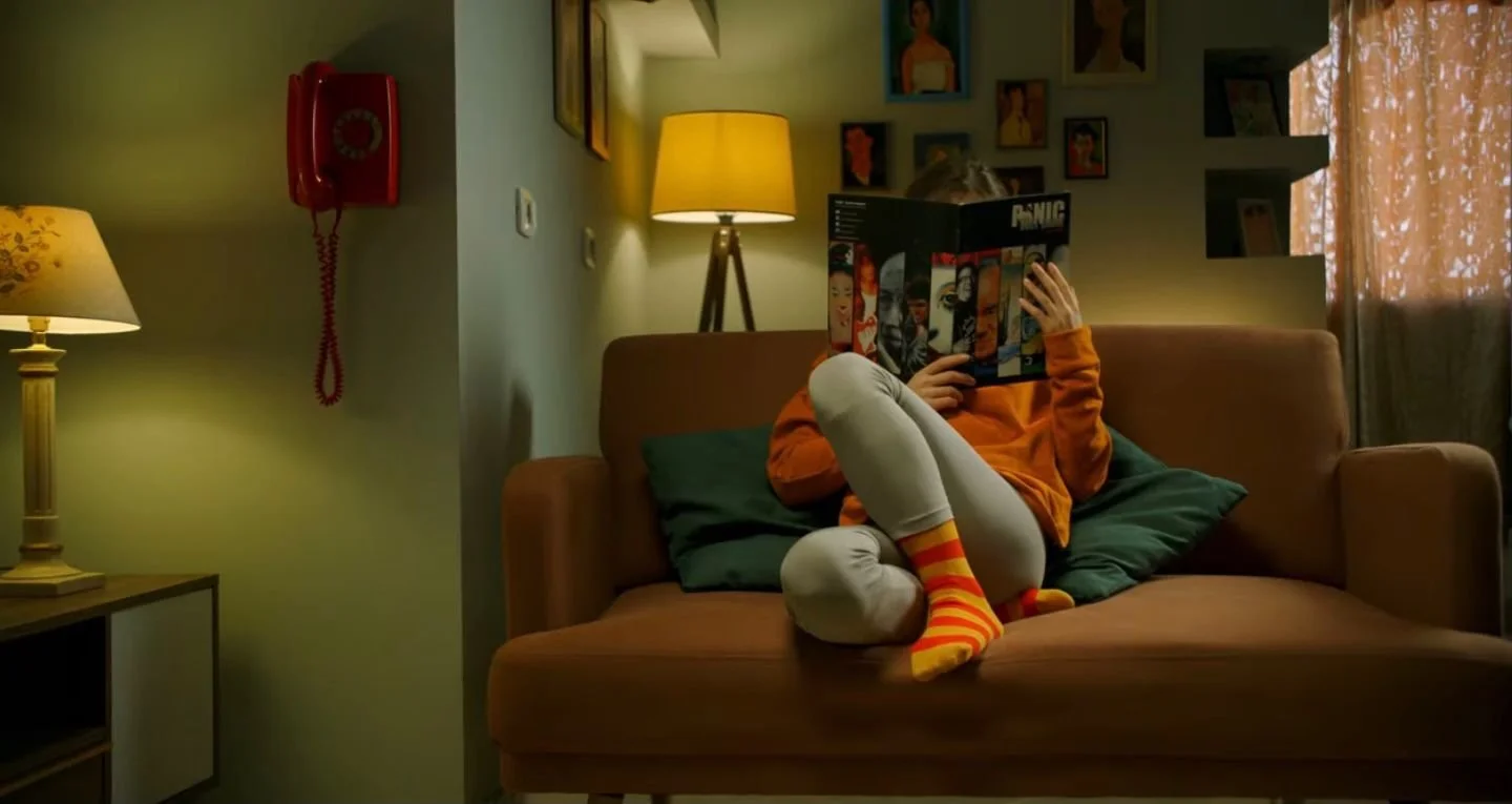 Person sitting on a brown couch reading a comic book, wearing orange sweatshirt, gray pants, and colorful striped socks, in a cozy living room with lamps and wall art.