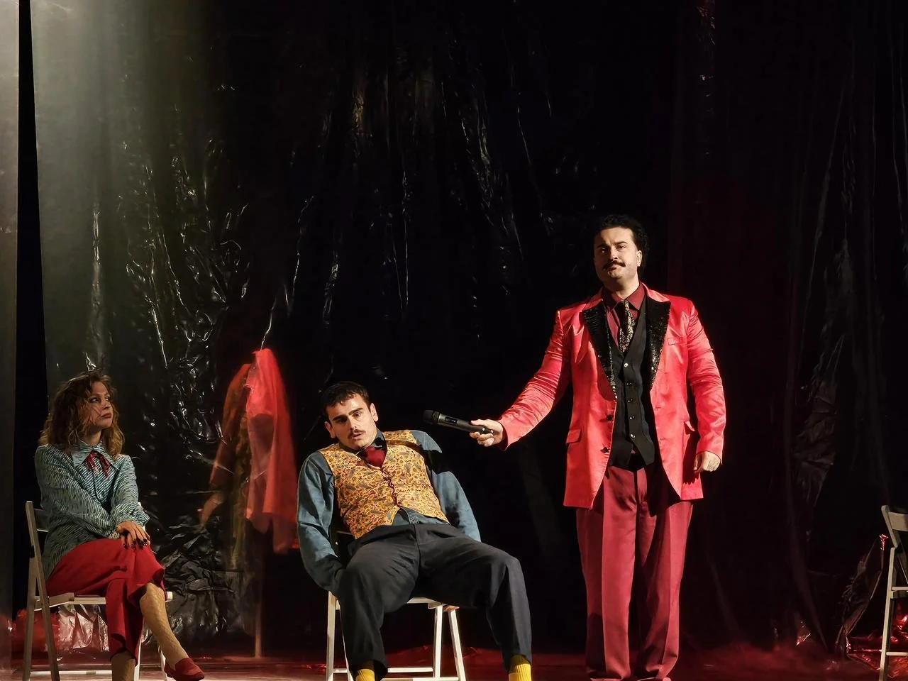 A theatrical performance with three actors on stage. A man in a bright pink suit holding a microphone, another man sitting in a chair with a gold vest and colorful shirt, and a woman sitting on a chair wearing a striped shirt and red pants.