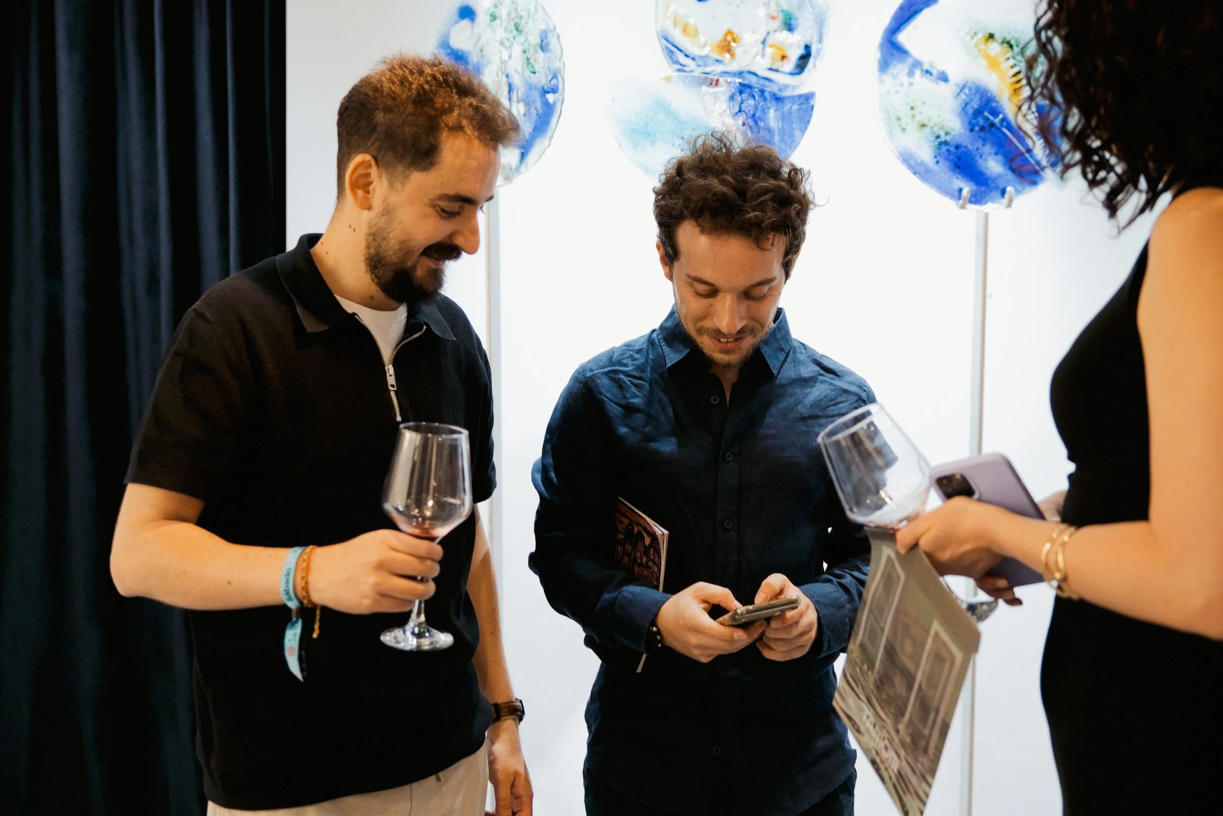 Three people are at a social event, chatting and looking at a smartphone. One person is holding a wine glass. There are blue and white decorative art pieces in the background.