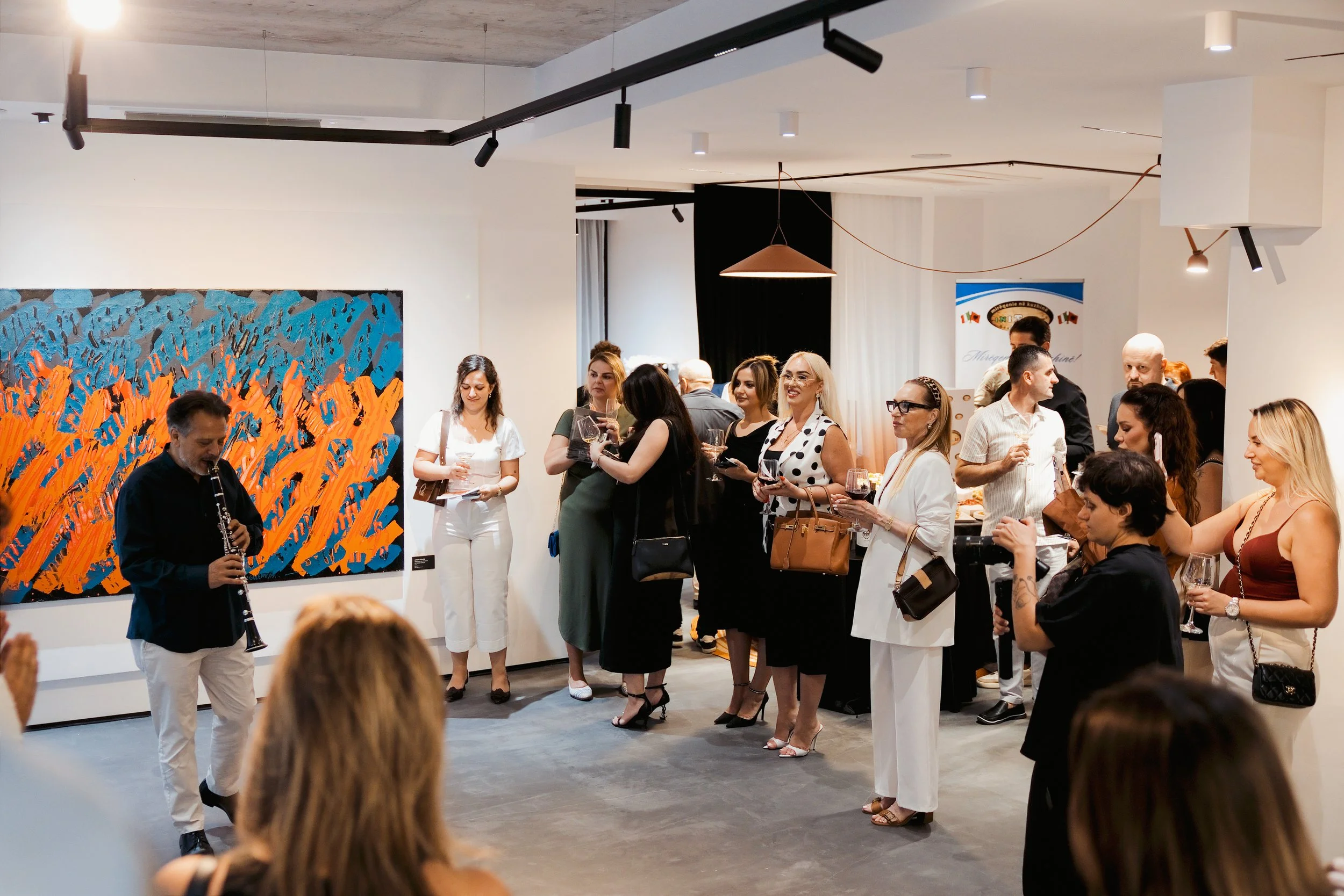 Group of people attending an art gallery event, standing and socializing while some look at artwork. A man is playing a clarinet. The gallery has white walls, modern lighting, and includes a large colorful abstract painting.