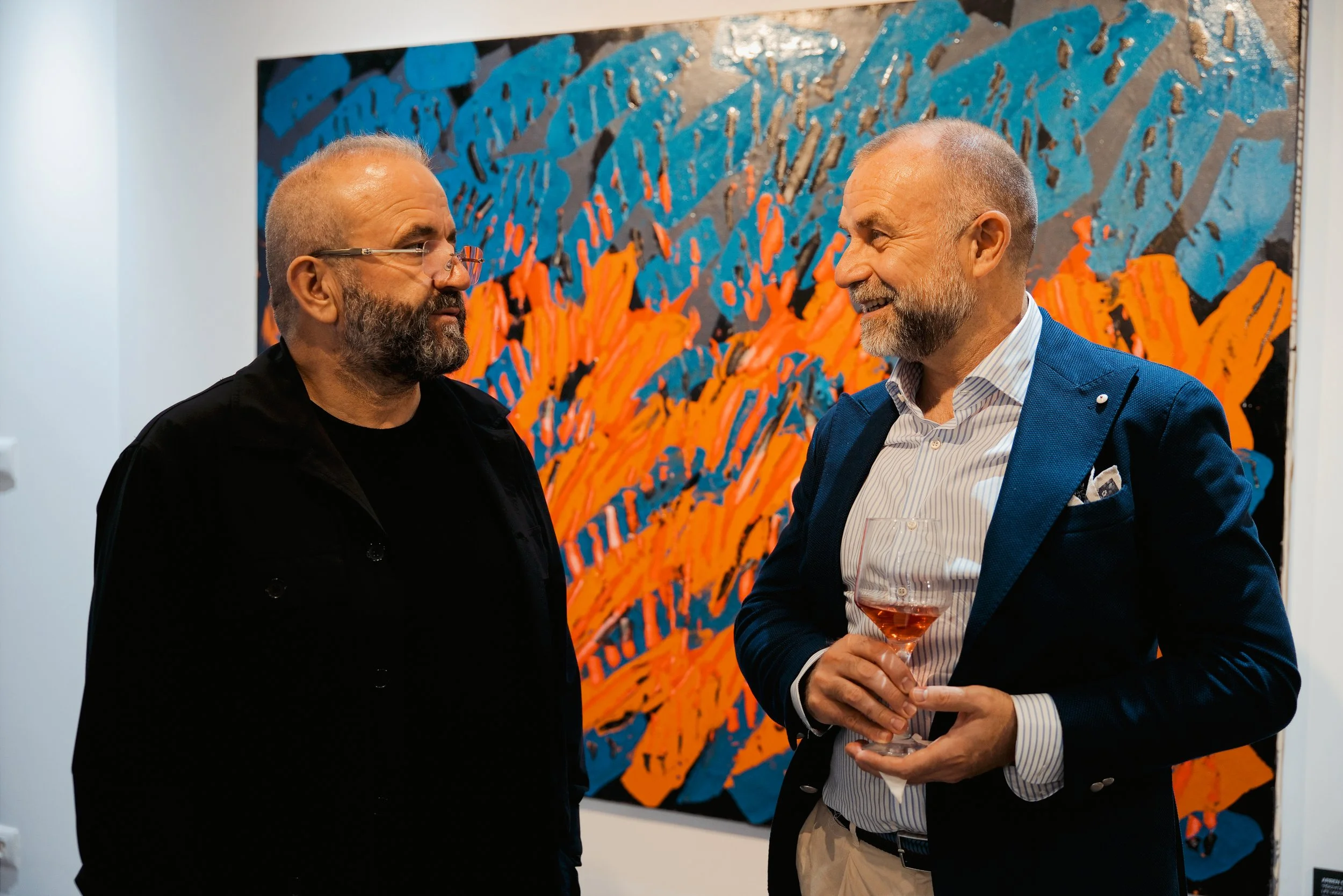 Two men having a conversation at an art gallery, with a colorful abstract painting of blue, orange, and black in the background. One man is holding a glass of rose wine.