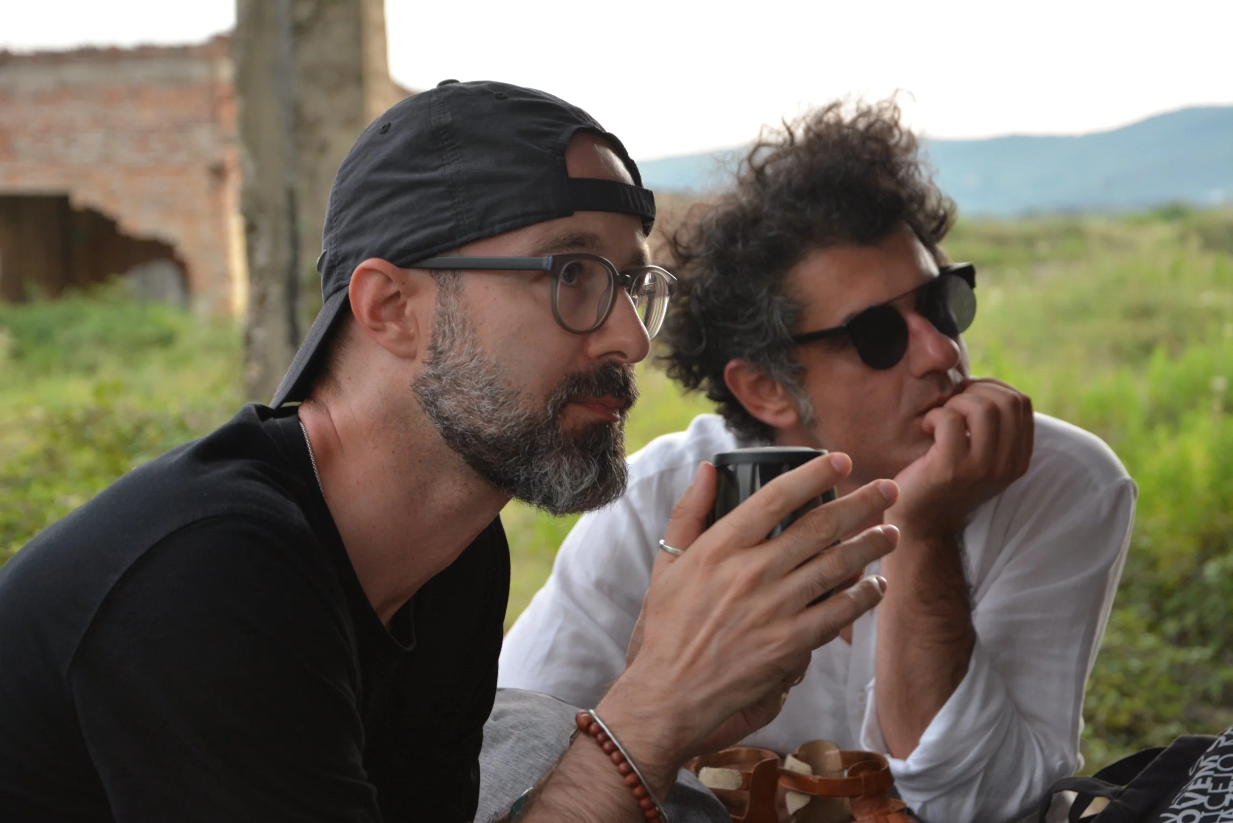 Two men sit outdoors, looking intently at a phone one is holding. The man on the left wears glasses, a black cap backward, and a dark shirt. The man on the right has curly dark hair, sunglasses, and a white shirt. In the background, there are green f