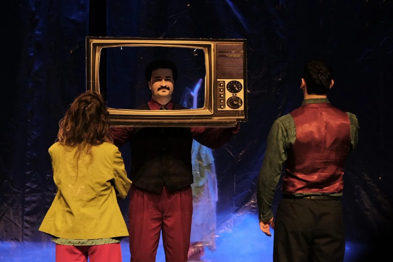 Three people on stage, one person holding a vintage television set in front of their face, displaying another person’s face on the screen, with a dark background and blue stage lighting.