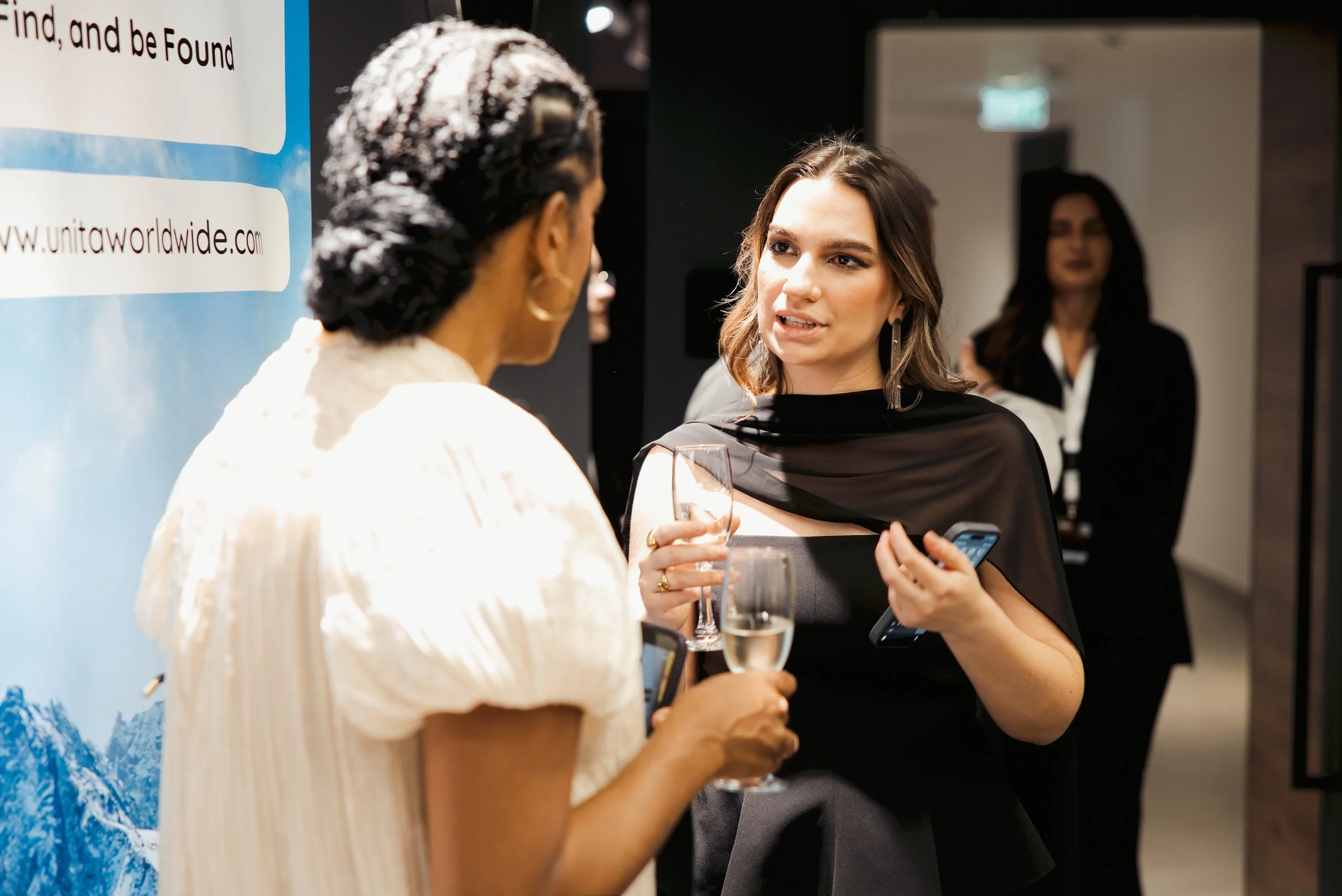 Two women are in conversation at a social event, one with dark hair and the other with curly hair, holding a glass of champagne, standing near a blue and white promotional banner.
