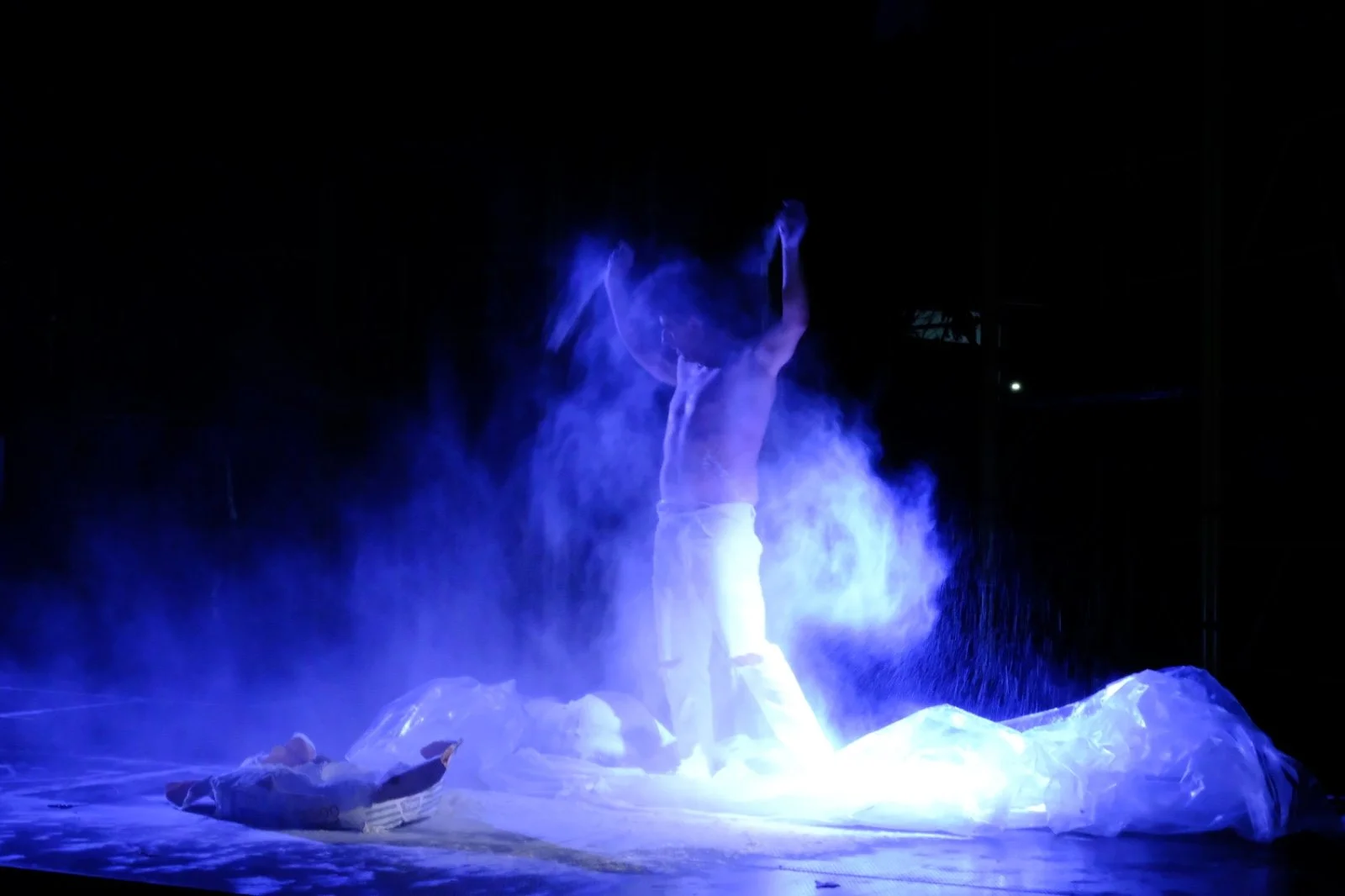 A performer dressed in white is on a dark stage, with dramatic blue lighting and smoke, performing a handstand or similar acrobatic move.