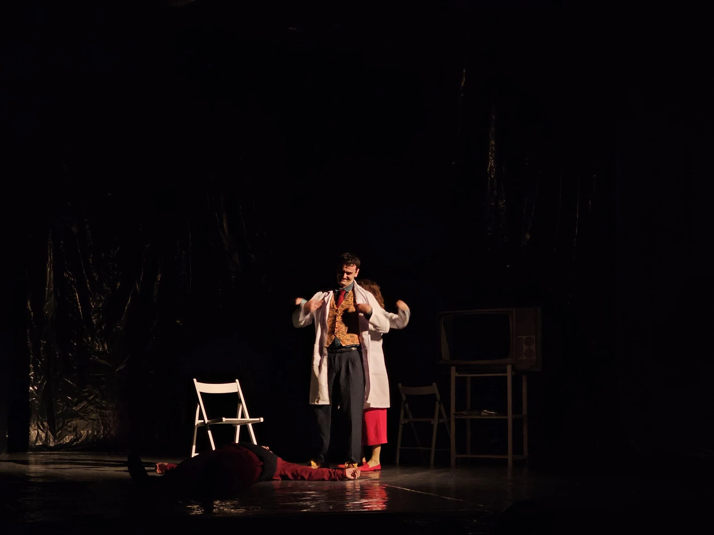 The image depicts a scene from a theatrical performance with two actors, one standing and one lying on the ground, in a dark stage setting with minimal props and chairs. The standing actor is dressed in a colorful outfit, possibly a nurse or doctor, 