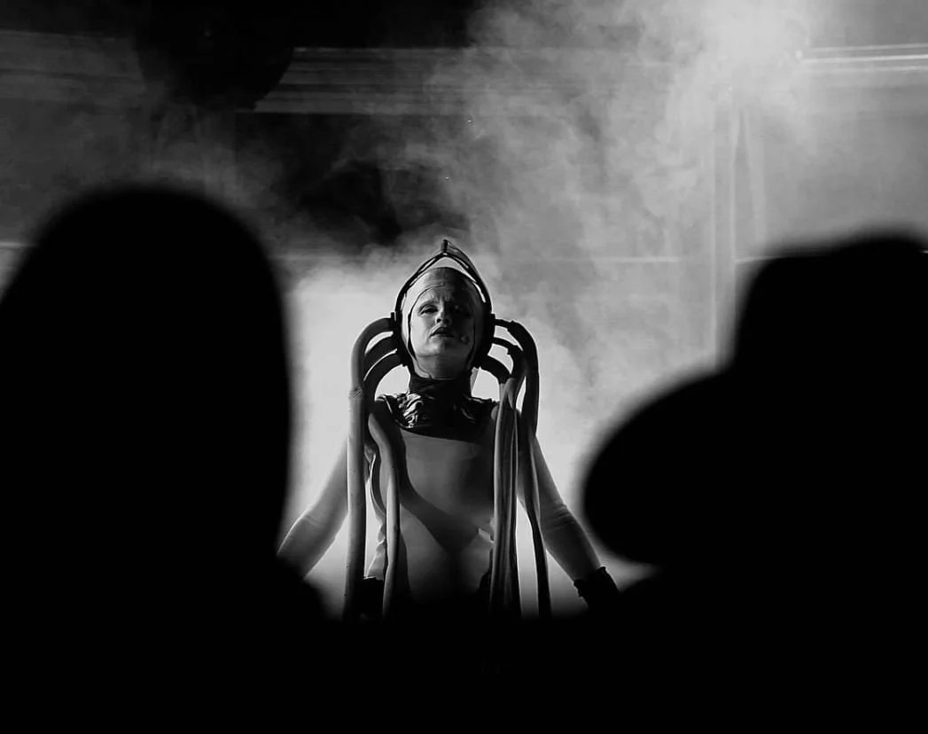 A person in a futuristic costume with a helmet and tubing, standing among silhouettes of audience at a performance or cosplay event.