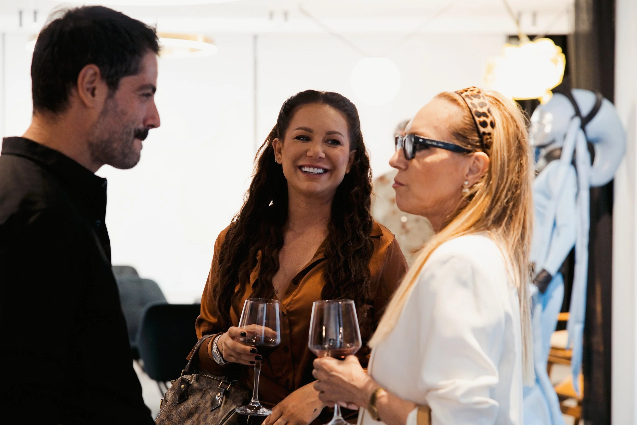 Three women and one man engaging in conversation at a social gathering, holding wine glasses.