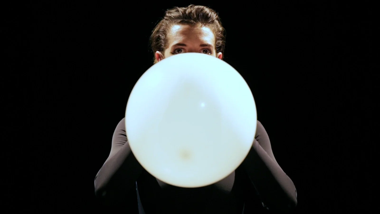 Person holding a glowing white balloon in front of their face, against a black background.