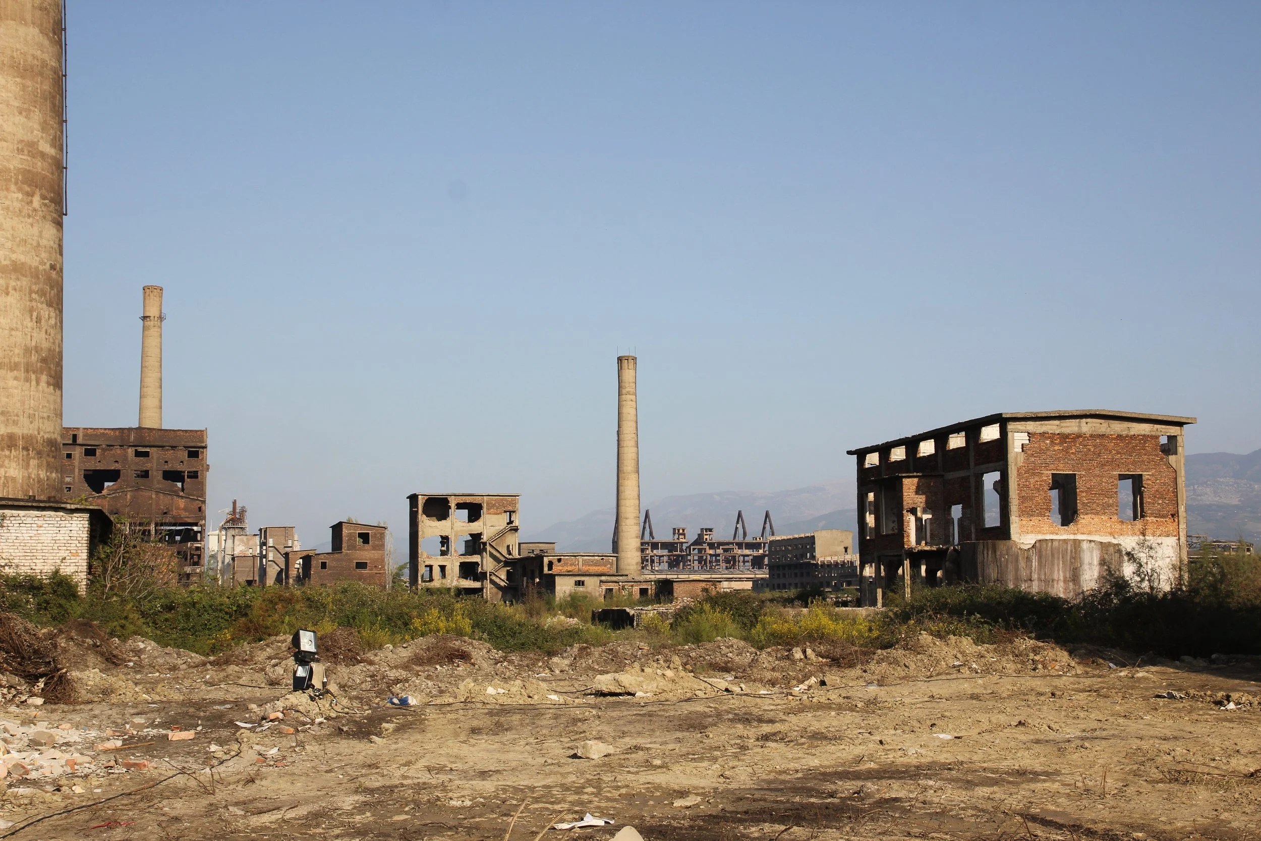 Ruined industrial buildings and towers in an abandoned area with overgrown vegetation and a dirt foreground.
