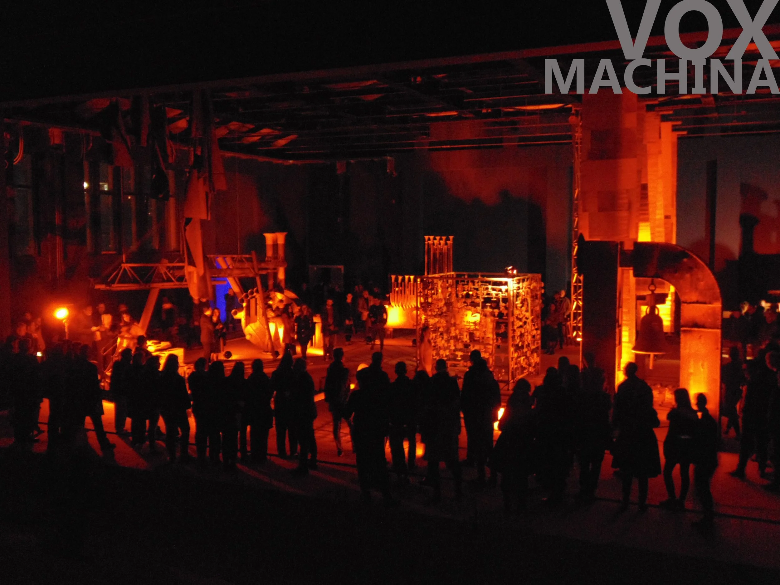 A dark indoor event space with orange lighting and silhouettes of people standing and watching a performance. Elements of stage or art installation are visible in the background.