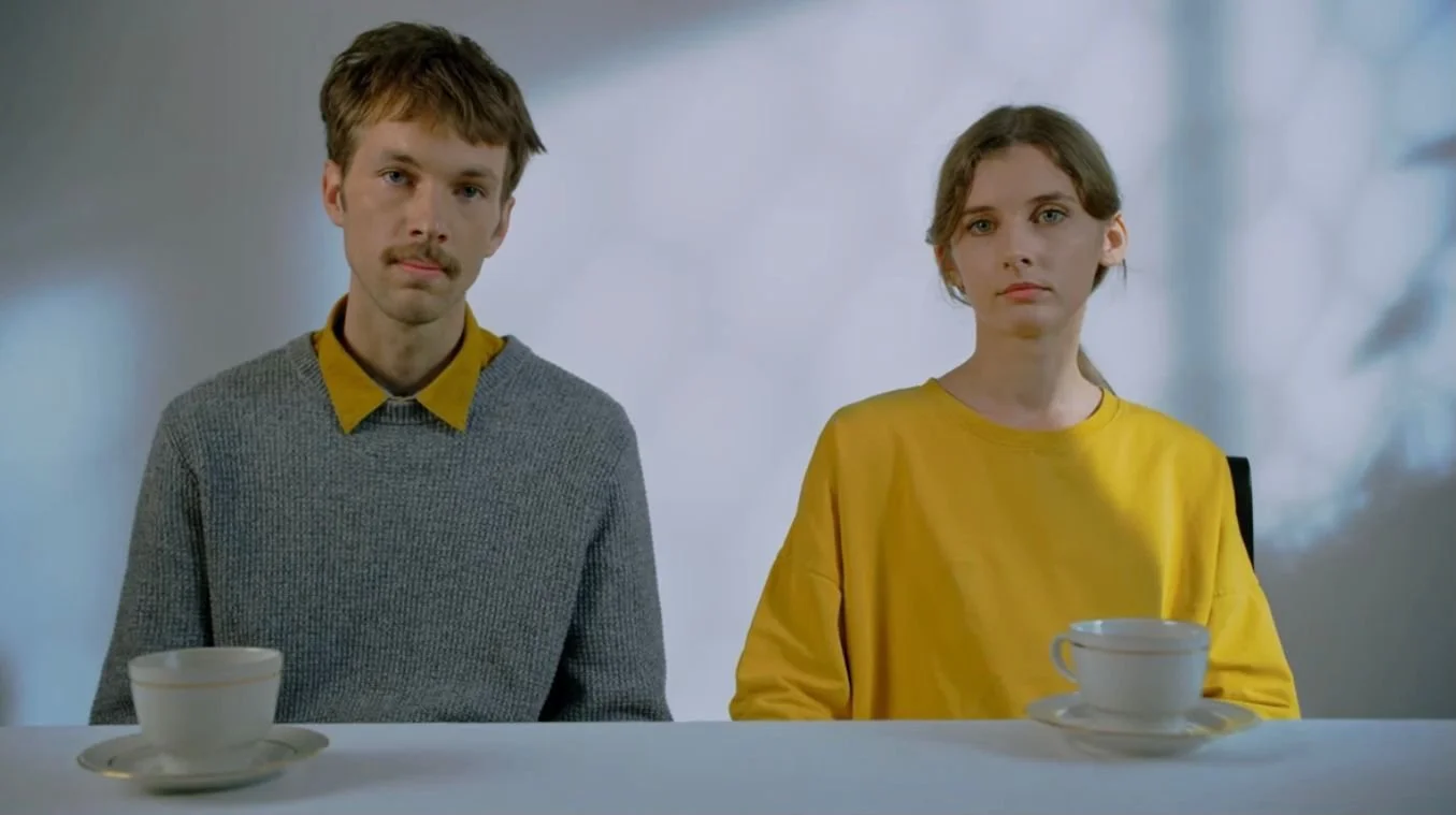 A man and woman sit at a table with cups in front of them, both looking directly at the camera with neutral expressions.