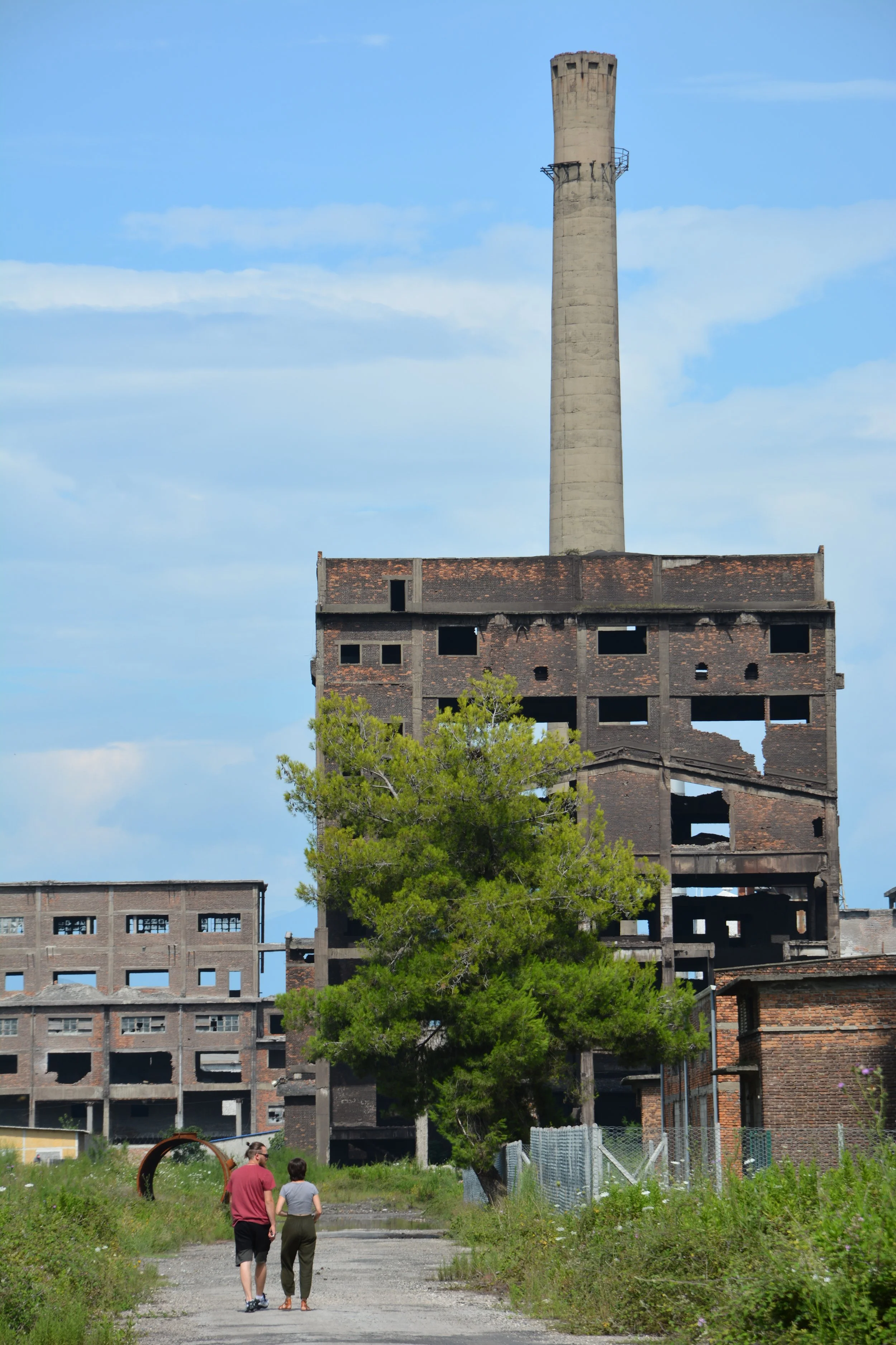 Two people walking on a gravel path surrounded by overgrown grass and bushes, with an old, abandoned brick building and a tall smokestack in the background, under a blue sky with some clouds.