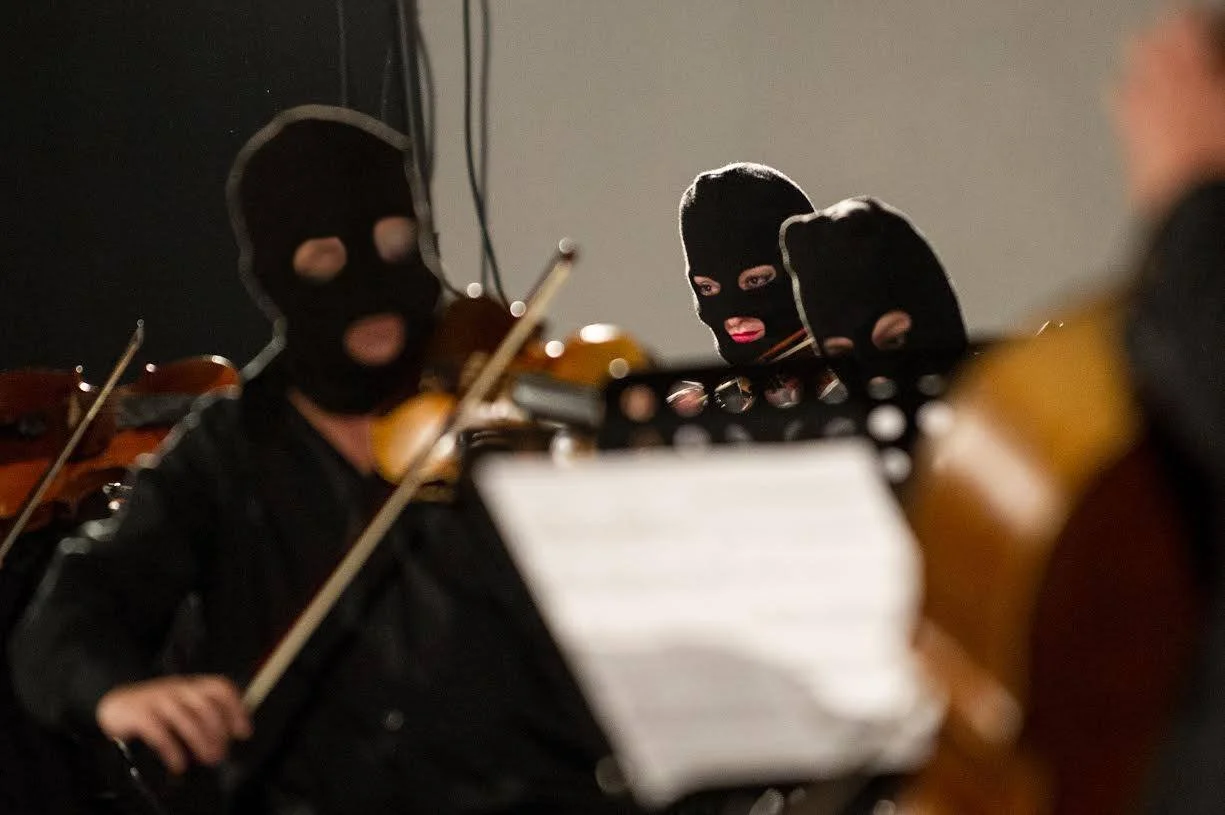 Three musicians wearing black ski masks playing violins in a dimly lit room.