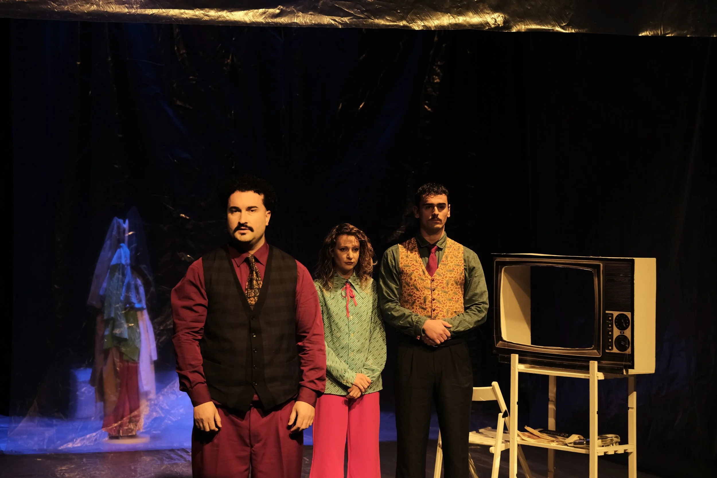 Three people stand on a stage in front of a dark background with theatrical props, including a vintage microphone and a white wooden table with chairs. The individuals are dressed in vintage-style clothing.