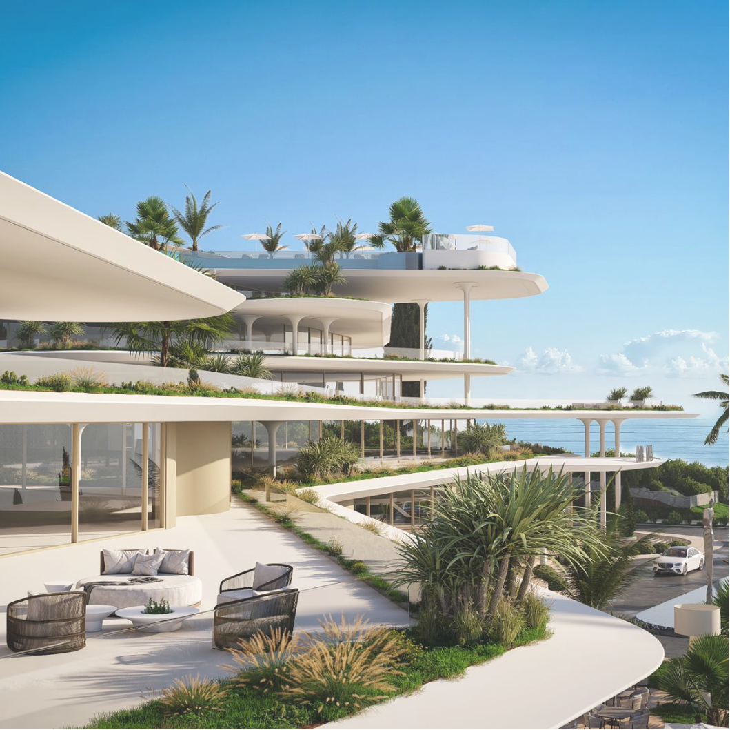 Modern multi-tiered beachfront building with white curving balconies, lush greenery, and palm trees, overlooking the ocean on a sunny day.