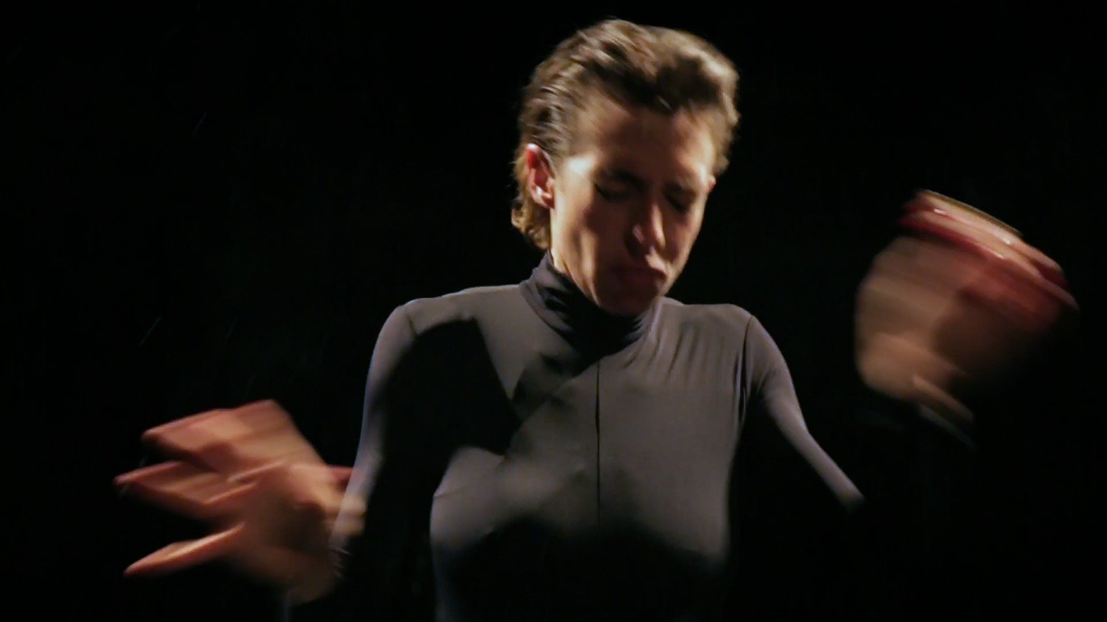 Person with short hair wearing a black high-neck shirt, moving with blurred hands in a dark setting.