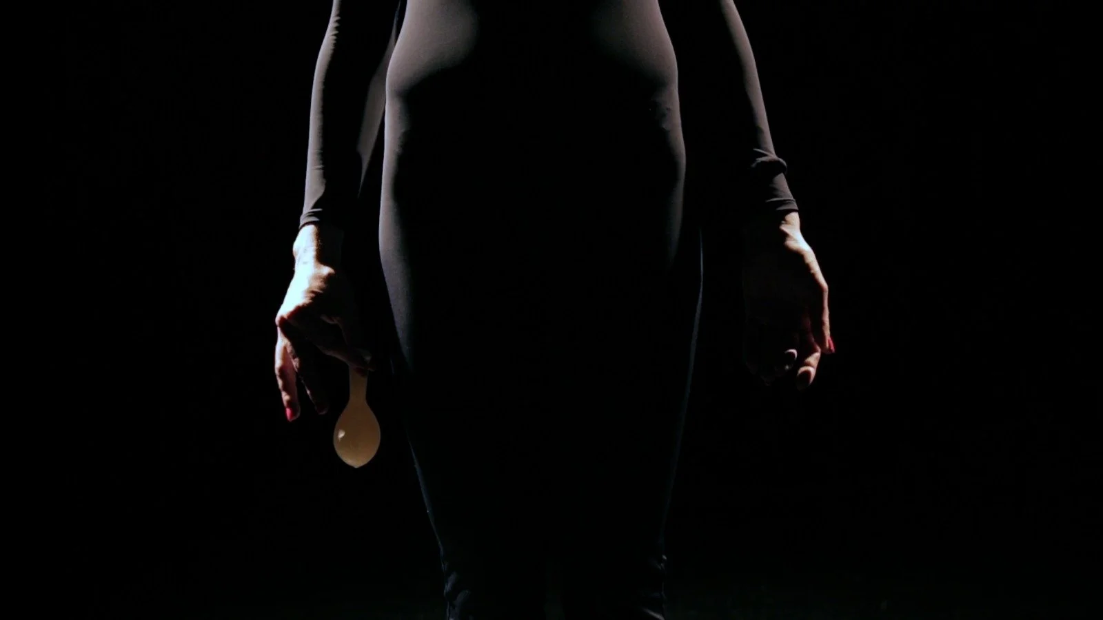 A person is standing in a dark setting, holding a masturbation device in one hand, with their body mostly obscured by shadows and high contrast lighting.