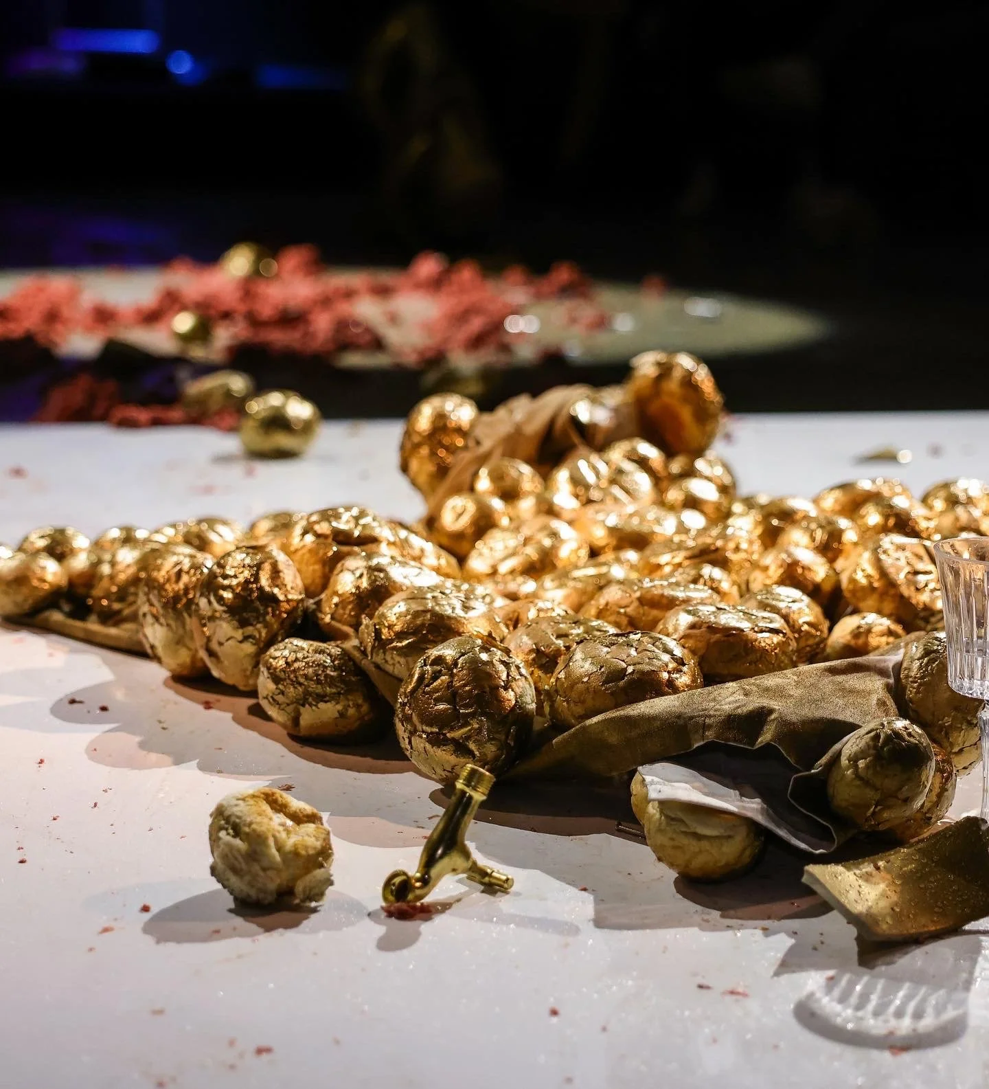 Gold-wrapped chocolates on a white table with a white background, decorated with a few gold ornaments and a small glass vase, blurred background with scattered chocolates and red decorations.