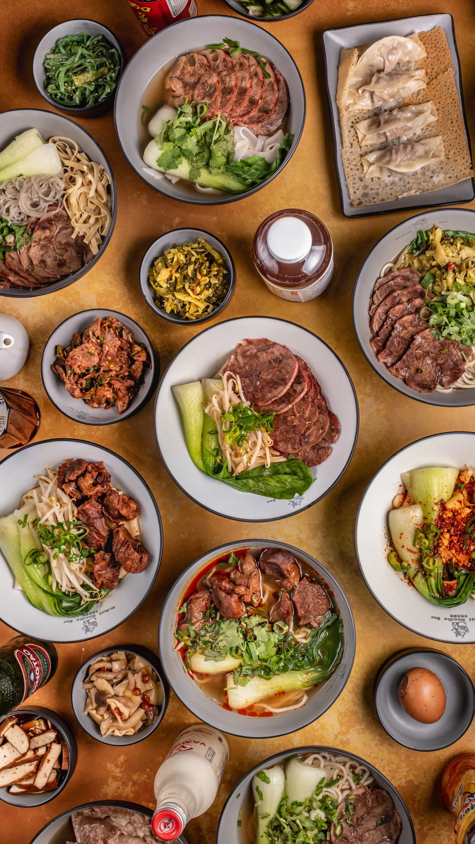 A variety of Chinese and Japanese dishes on a table, including bowls of noodles, steamed vegetables, dumplings, sliced meats, pickled vegetables, and beverages.