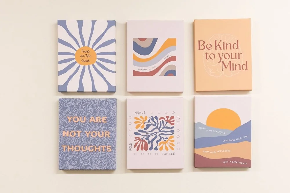Six colorful motivational and self-care quote cards arranged in two rows on a light wall. Top row: a card with blue and white radial patterns and a yellow circle, another with abstract shapes and the text "Healing is not" visible, and a peach-colored card saying "Be Kind to your Mind." Bottom row: a blue card with intricate patterns stating "You are not your thoughts," a white card with leaf illustrations and the words "Inhale" and "Exhale," and a card with stylized mountains, sun, and words like "Relax your forehead," "Unclench your jaw," "Drop your shoulders," and "Take a deep breath."
