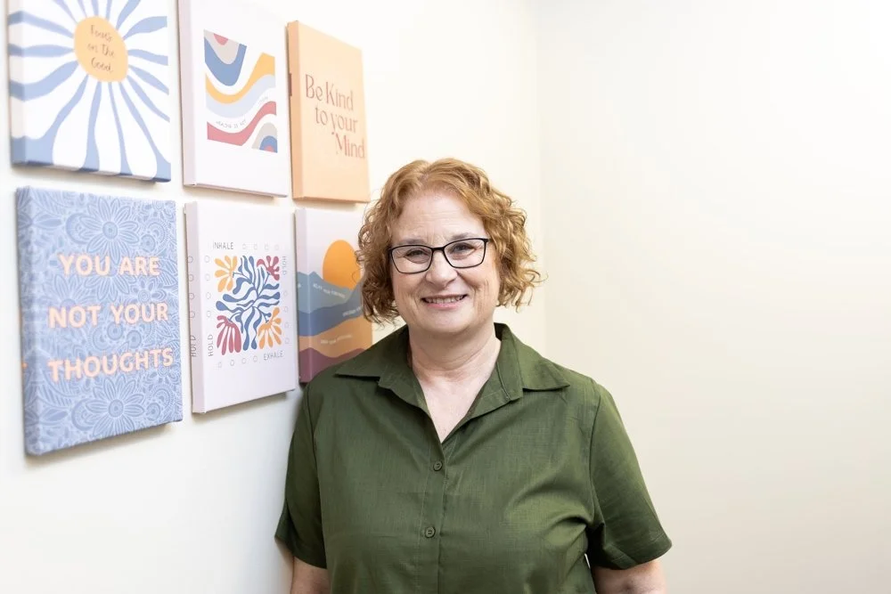 Karen Feeney LICSW, wearing glasses and a green shirt, standing in front of a wall with colorful inspirational quotes and decorations.