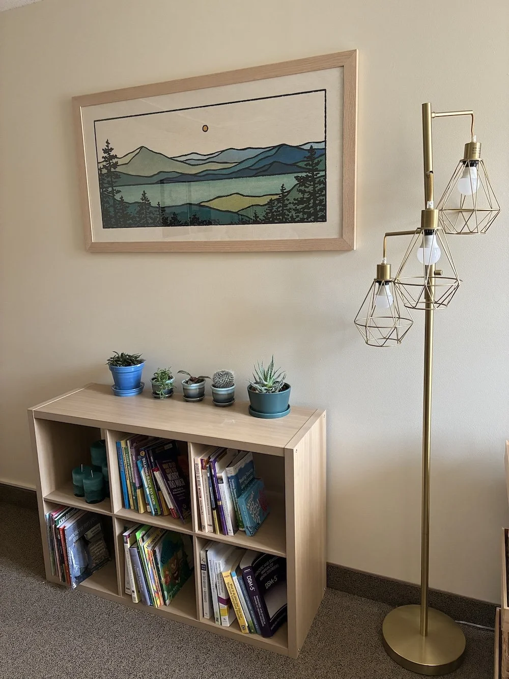 A wooden bookshelf with books and potted plants on top, a tall gold floor lamp with geometric light covers, and a landscape artwork with mountains and trees hanging on the wall.