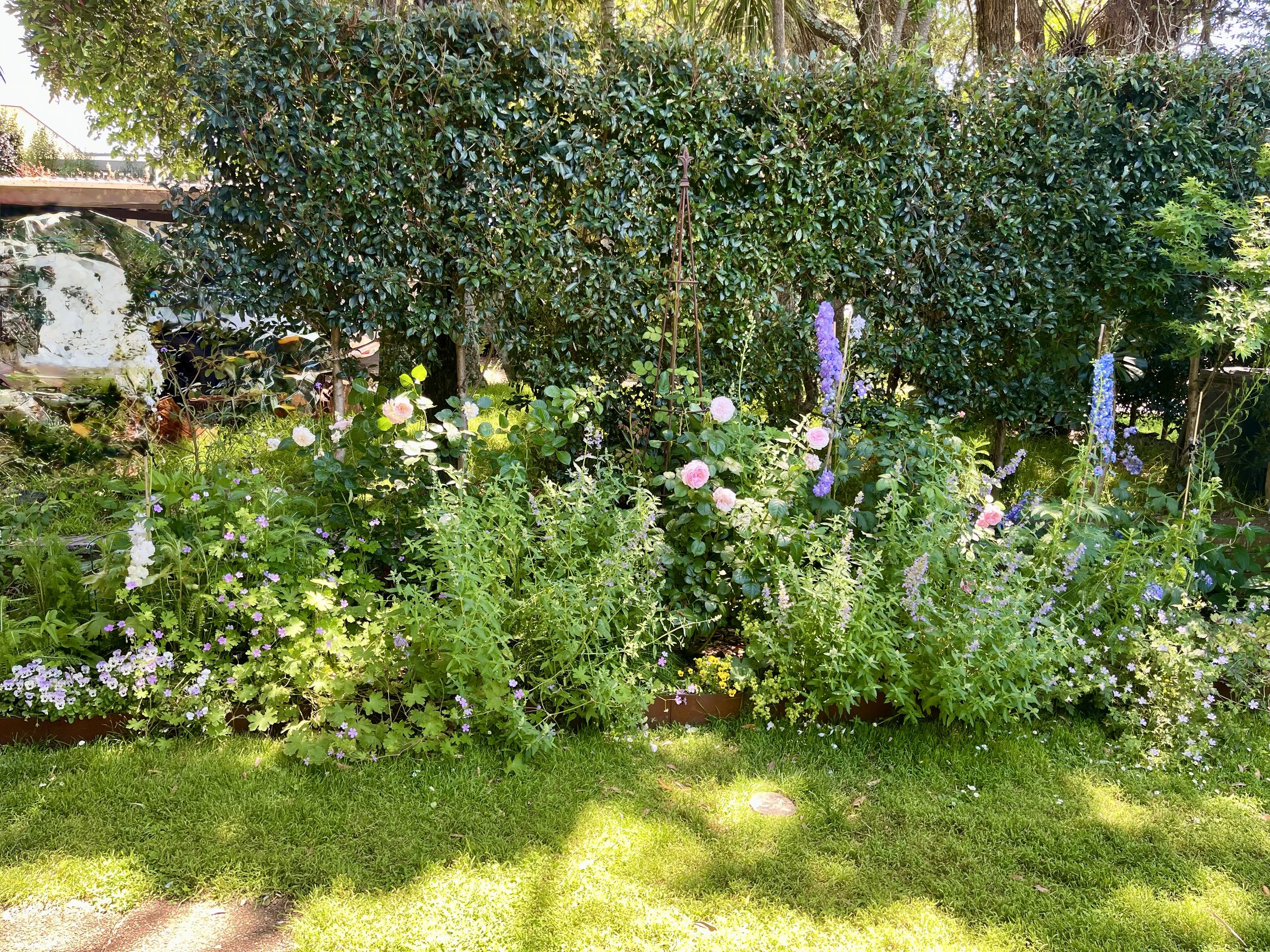 A lush garden with a variety of flowering plants, a green lawn, and a dense hedge. Tall trees are visible in the background, and a small garden ornament is hanging from the hedge.