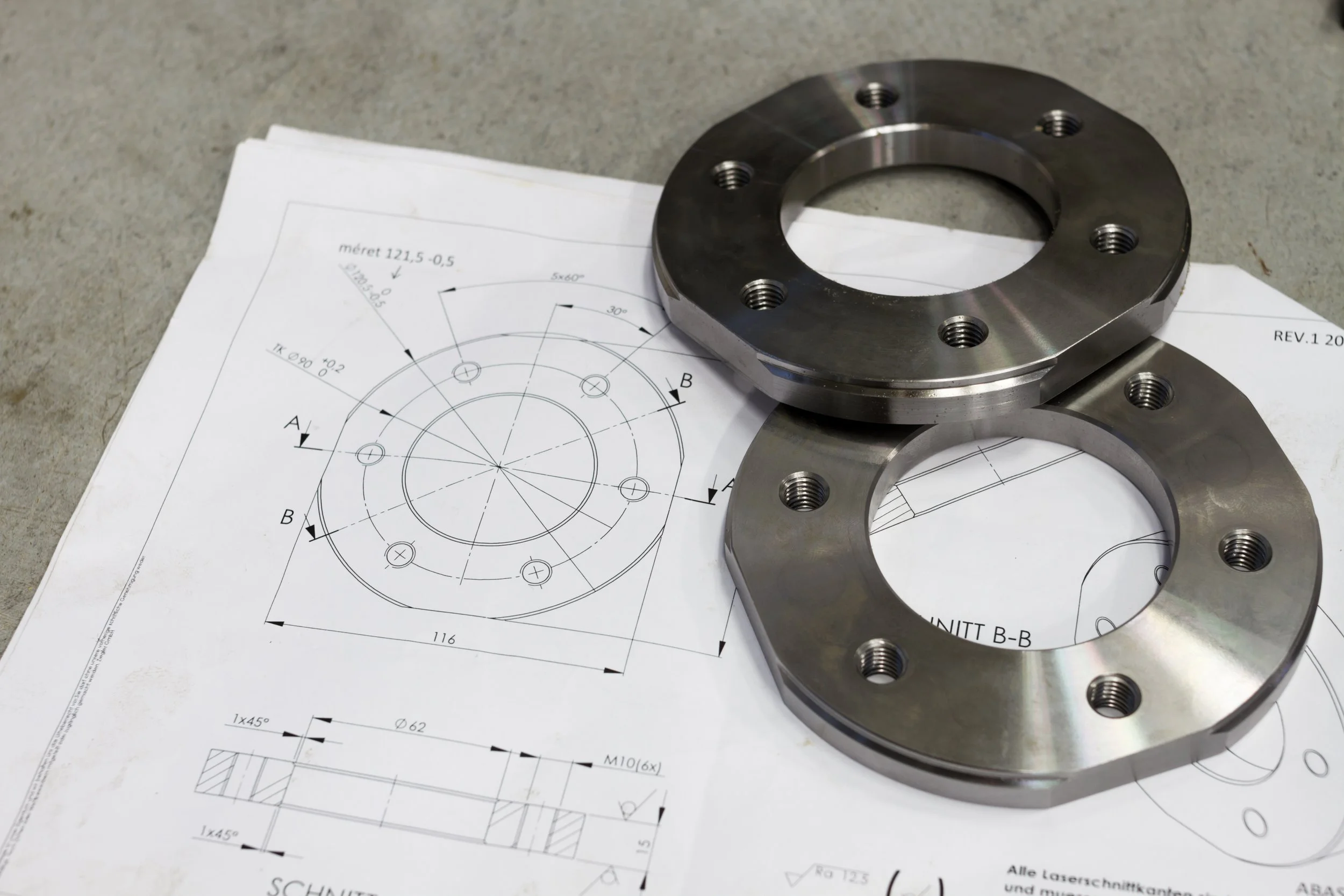 Two metallic circular mechanical parts with multiple holes on top of technical engineering drawings on paper.