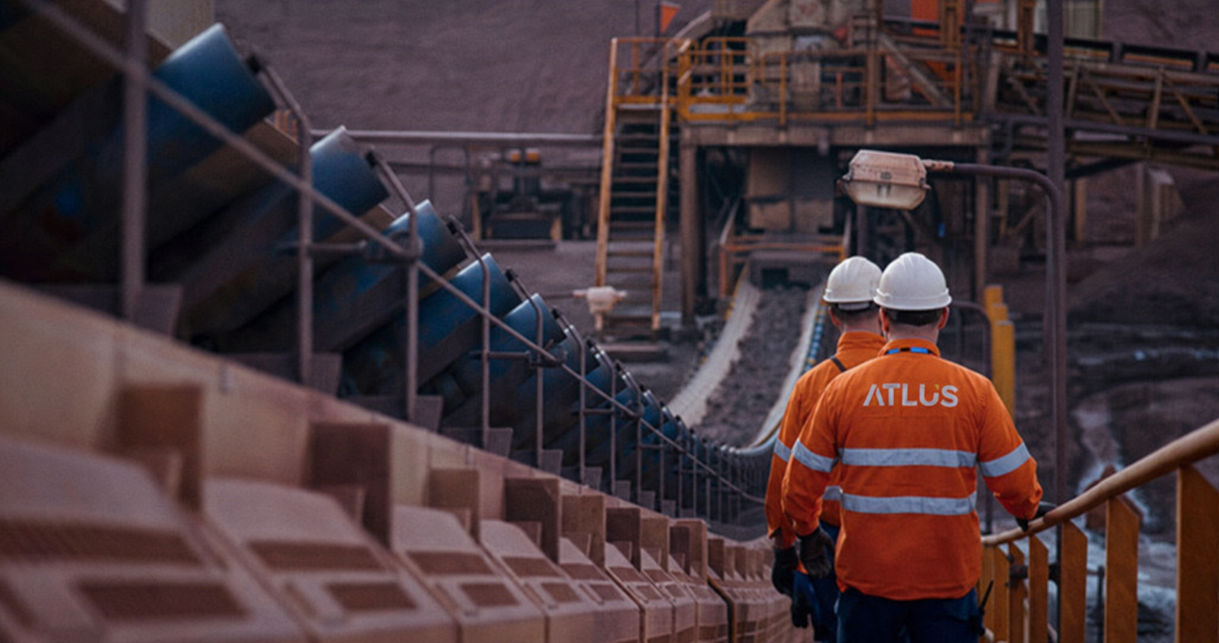 Two workers in orange safety jackets and white hard hats walking inside an industrial mining site with equipment, stairs, and conveyor belts visible.