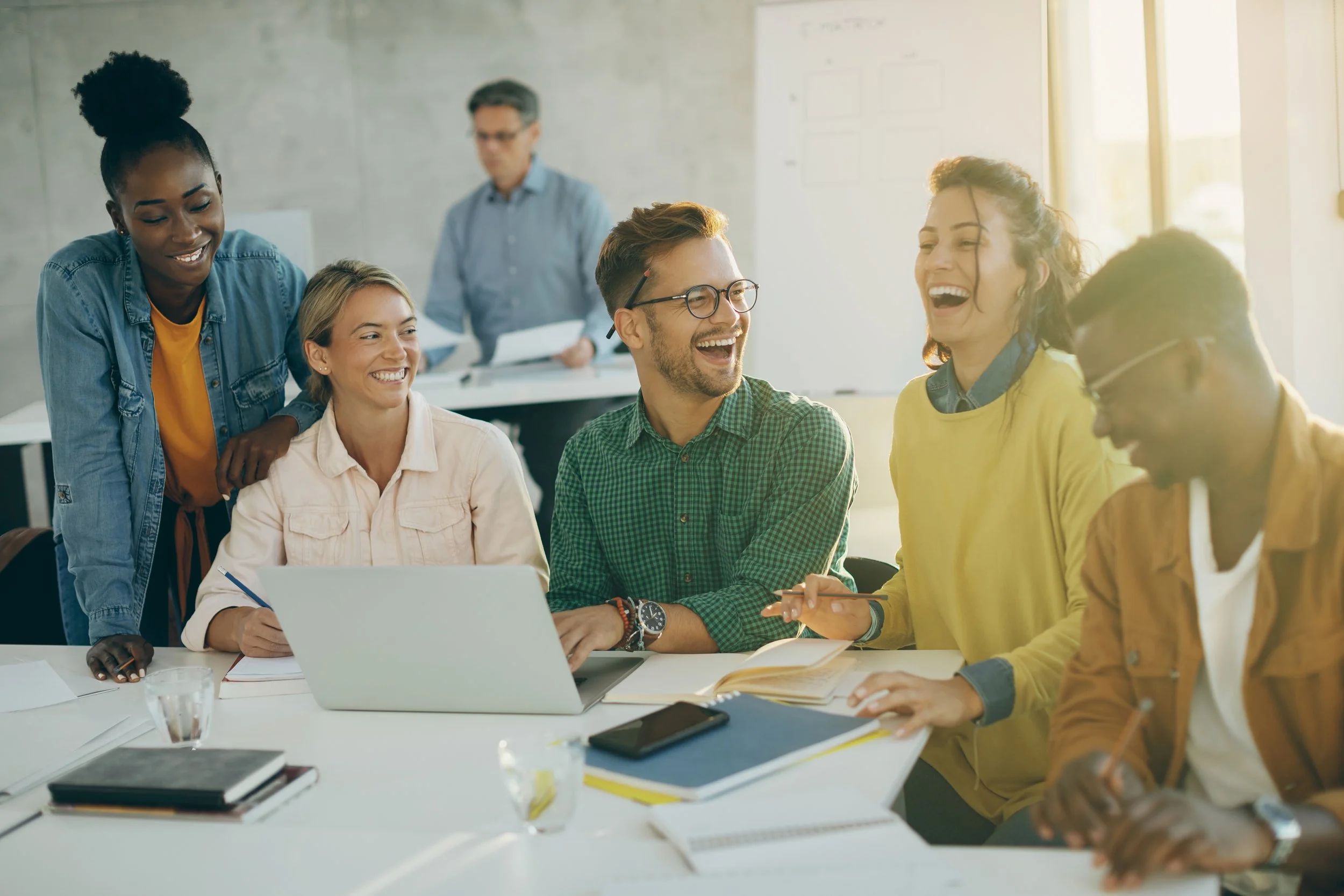 A diverse group of colleagues in a meeting room, smiling and laughing, with laptops and notebooks on the table, sunlight coming through large windows.