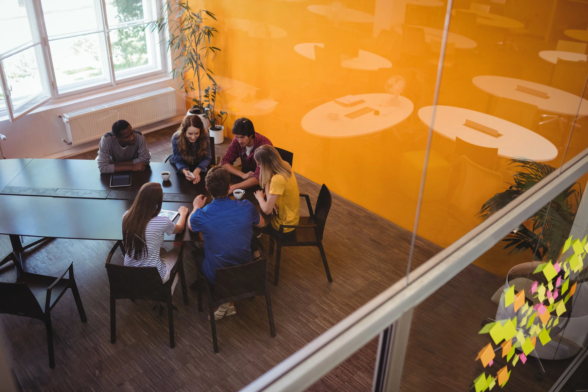 A group of six young adults sitting around a black table in a brightly lit room with large windows and a yellow wall. Some of them have coffee cups and a tablet on the table, and they are engaged in a discussion.