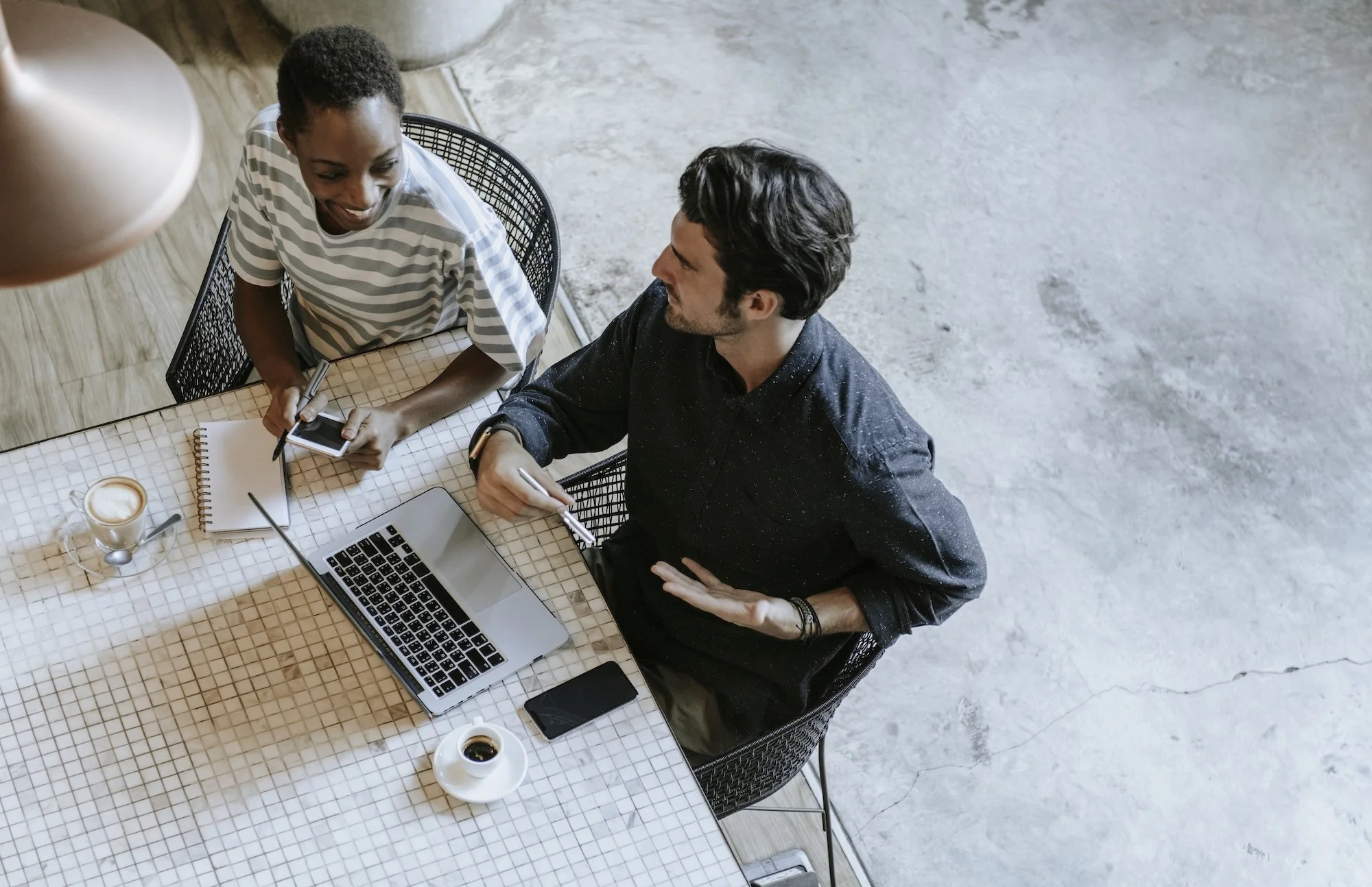 A woman with short curly hair and a striped shirt sitting at a table talking to a man with dark wavy hair and a dark button-up shirt. There is a laptop, smartphone, coffee, and notepad on the table.