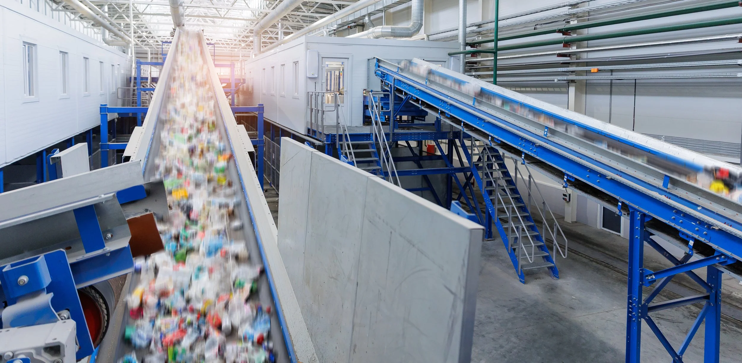 Industrial recycling facility with conveyor belts sorting and moving recyclable materials. Bright lighting and industrial piping visible.