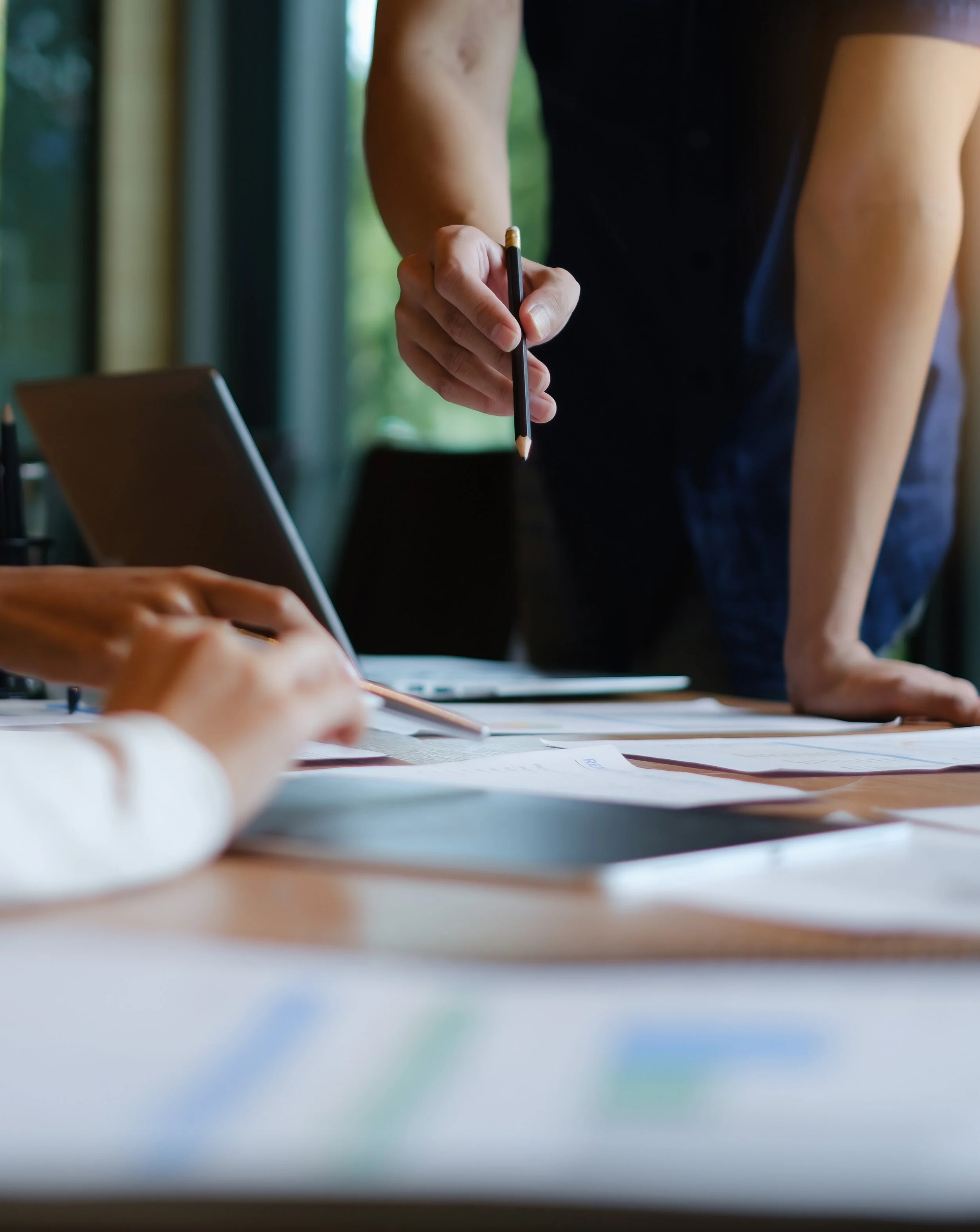 Person holding a black pencil over papers on a desk during a meeting or discussion.