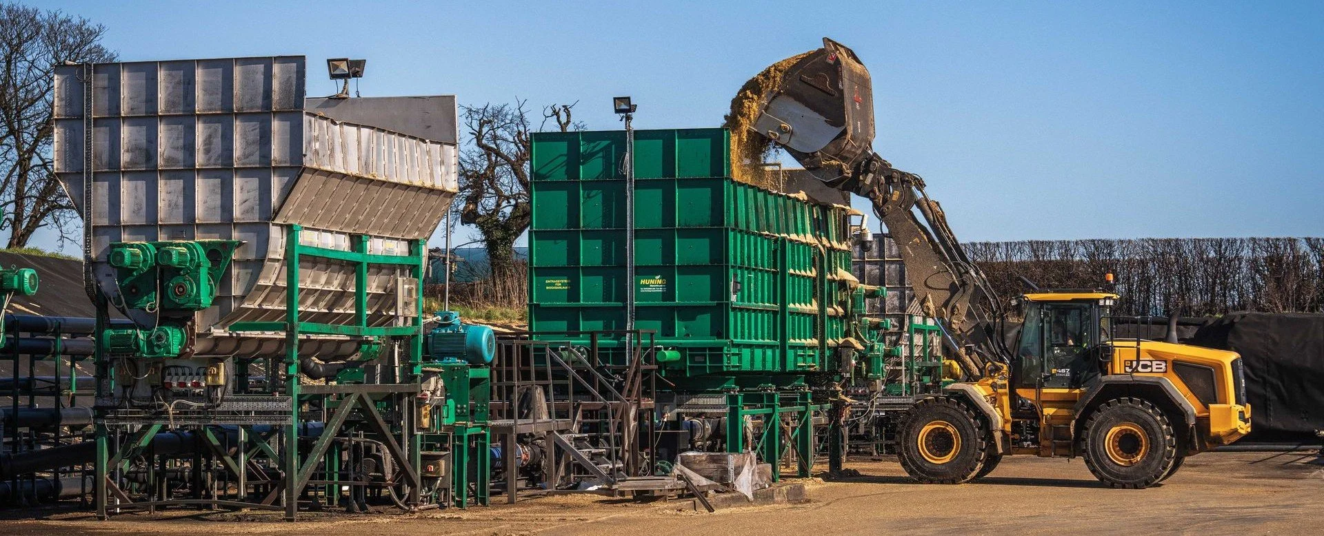 A large yellow JCB front loader with a mechanical arm blends materials into a green recycling container at an outdoor industrial site, with a clear blue sky and trees in the background.