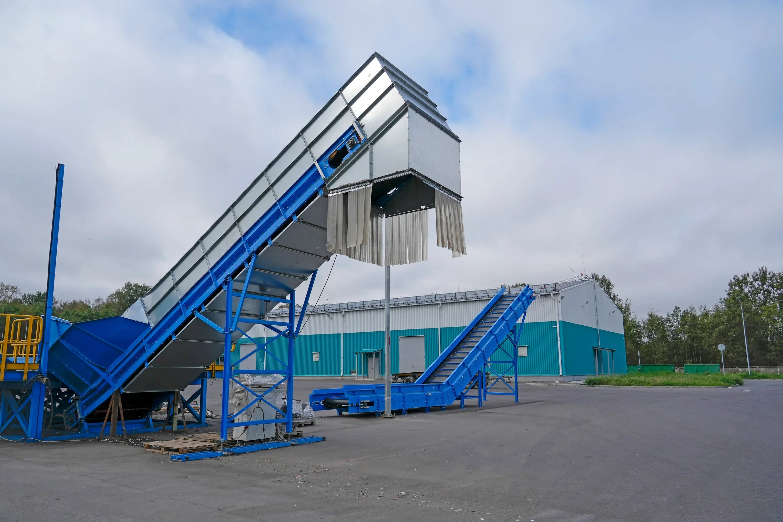 Large blue industrial machine with conveyor belts in an outdoor lot, with a blue and white building and cloudy sky in the background.