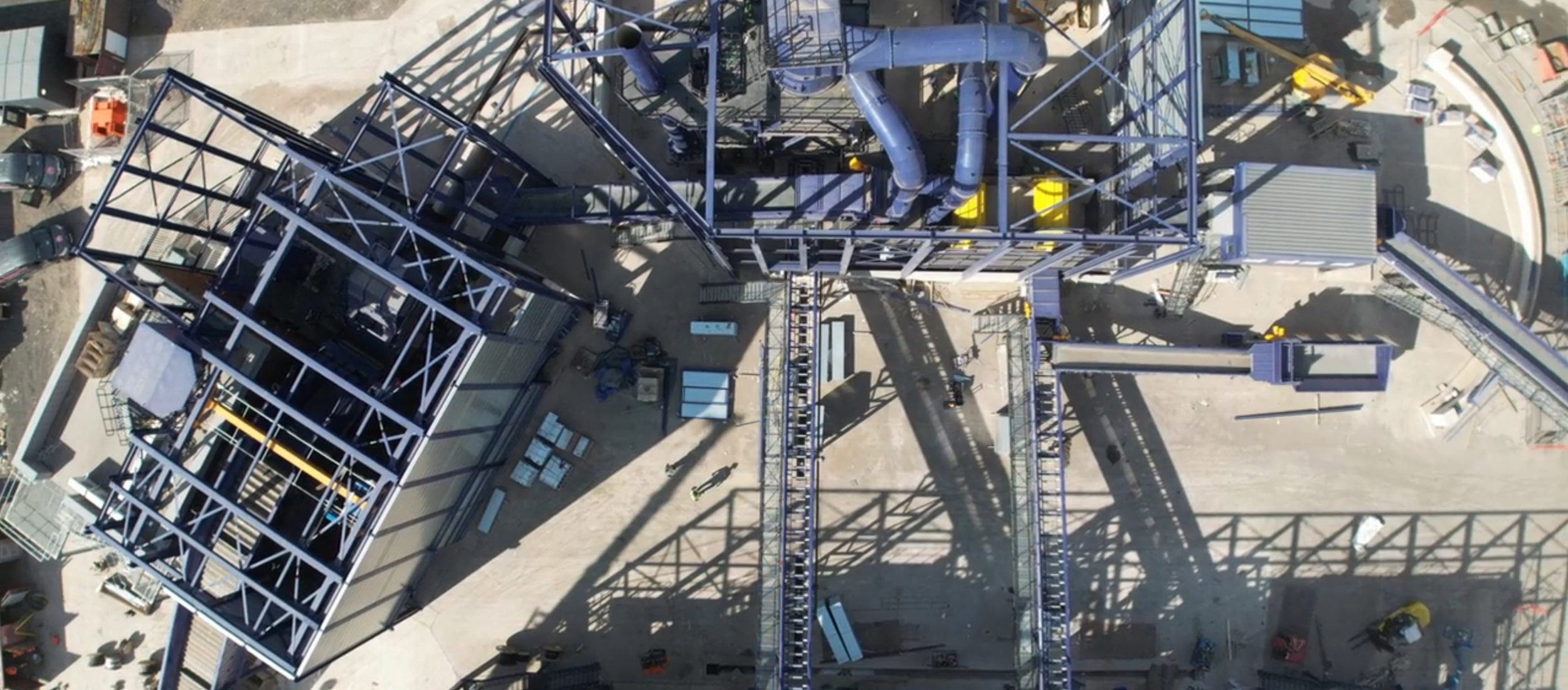 An aerial view of a construction site with steel framework, scaffolding, and construction workers. There are parked cars, equipment, and supplies around the site.